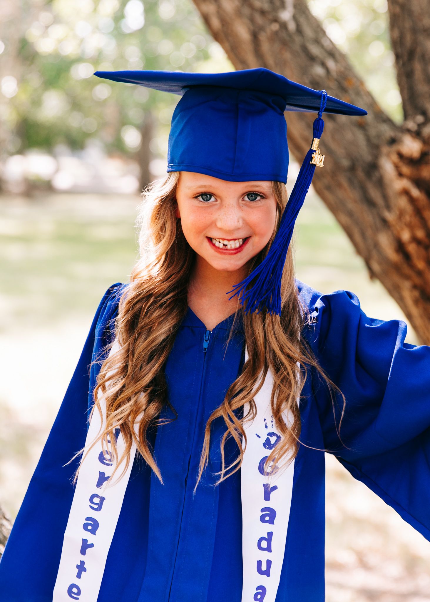 Smiling young girl in blue kindergarten graduation cap and gown outdoors.