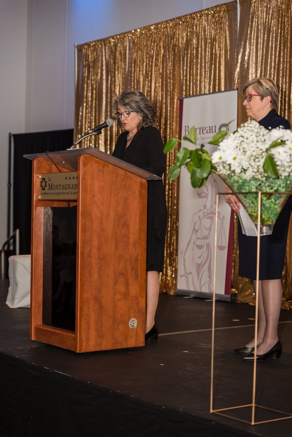 Speaker presenting at wooden podium with gold sequin backdrop and floral arrangements.