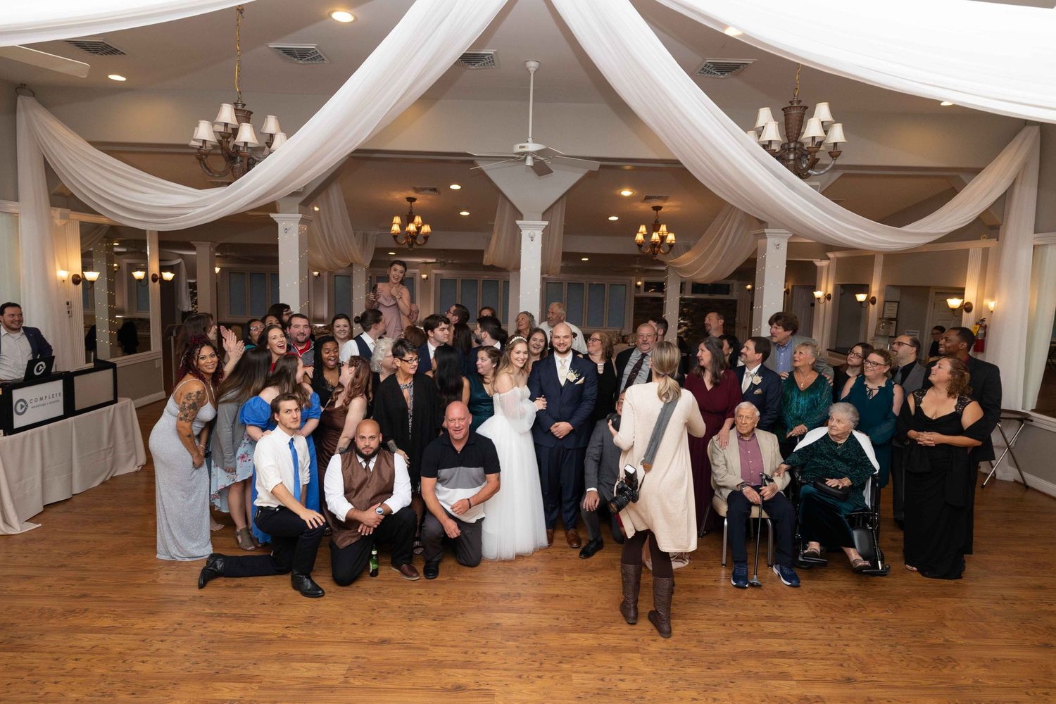 Large group wedding photo in decorated reception hall with white draped ceiling and chandeliers.