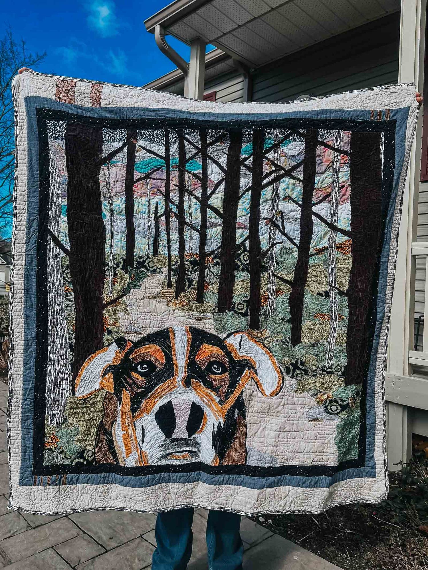 Art quilt showing a dog in the foreground and a forest in the background.
