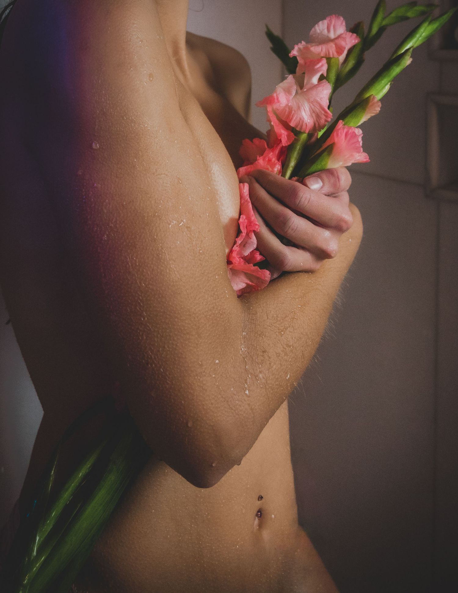 Close-up of a woman holding pink gladiolus flowers against her bare torso with water droplets.