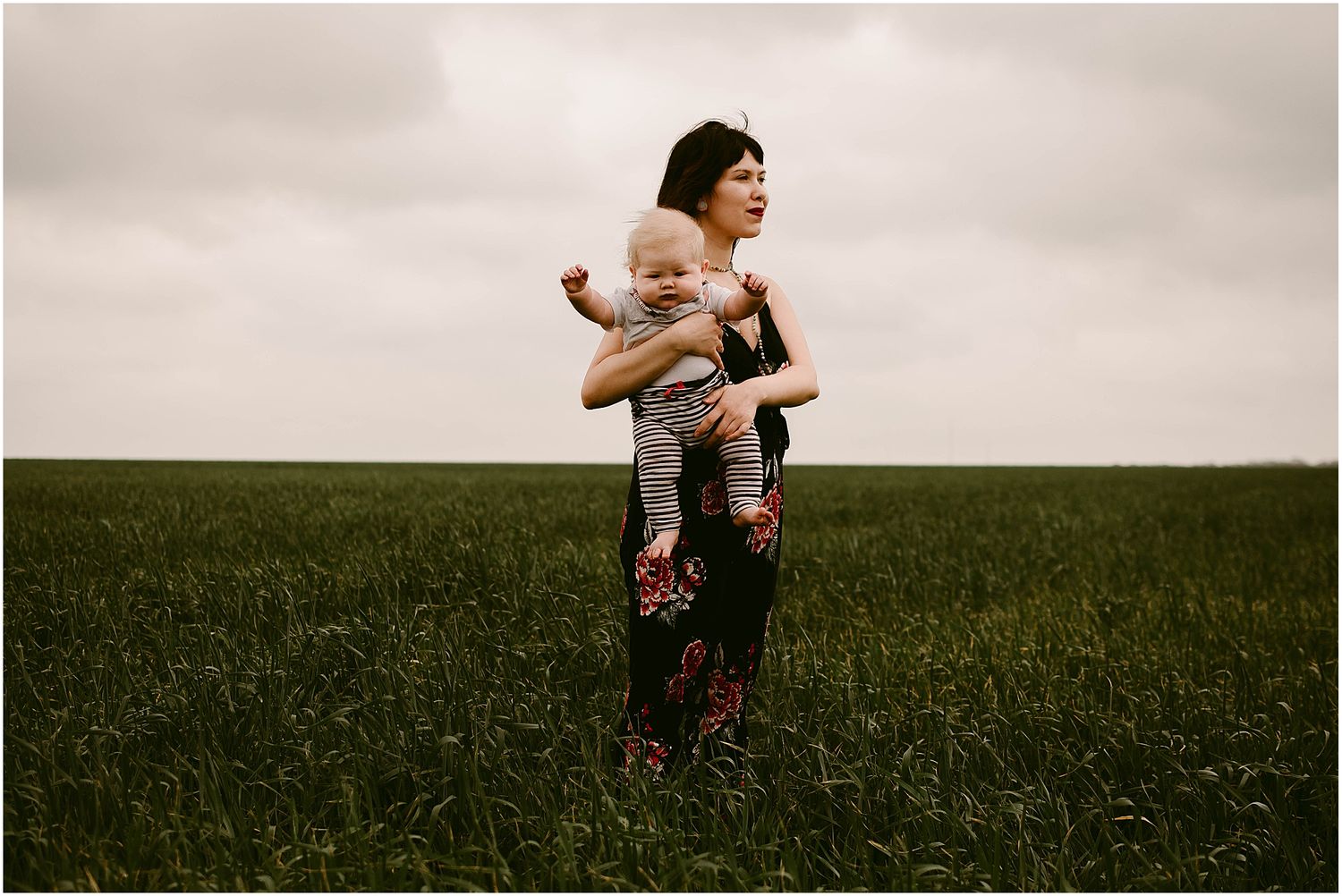 Adult holding infant stands in a misty field wearing floral pants and dark top.