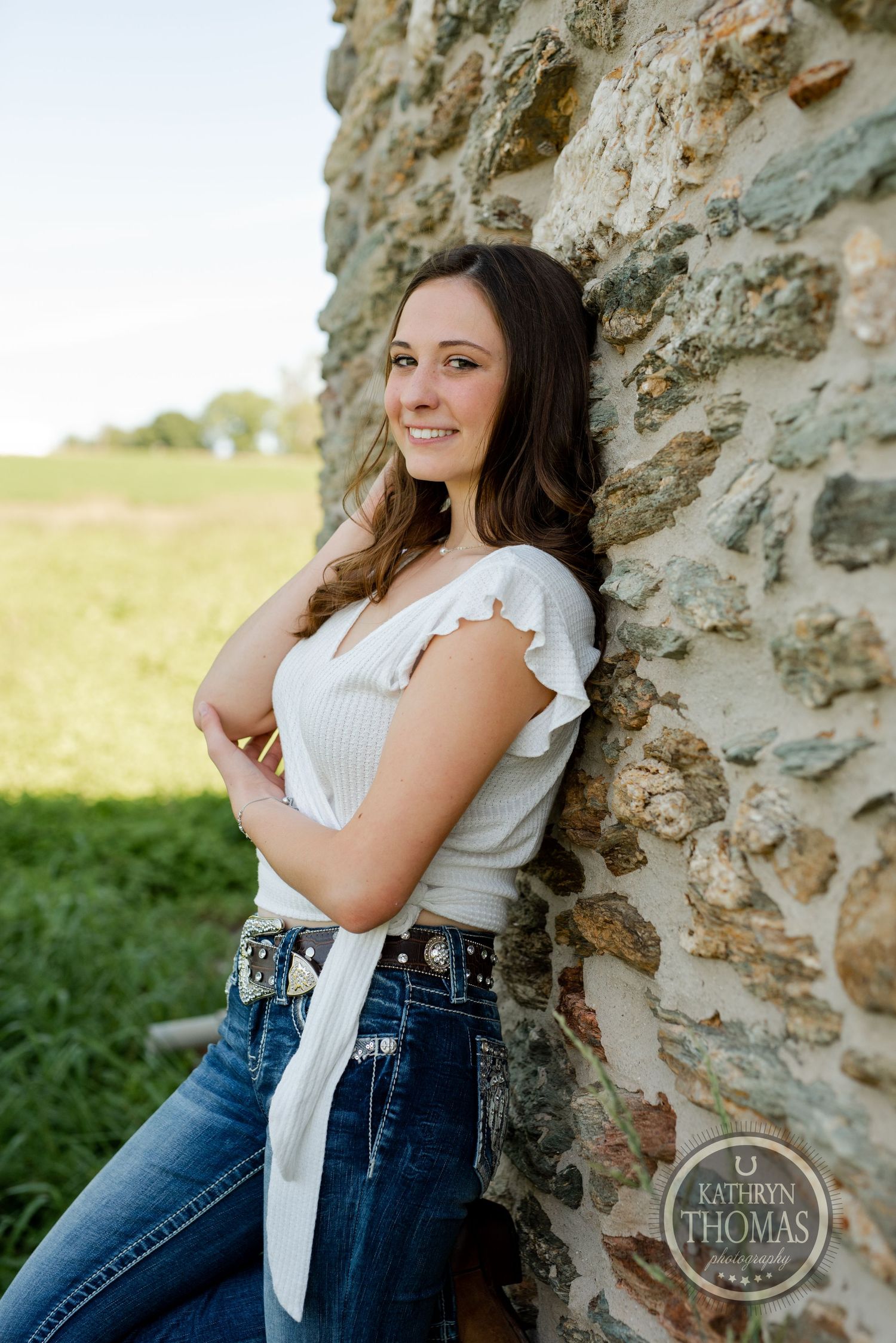 A woman in casual attire leans against a stone wall in natural outdoor lighting.