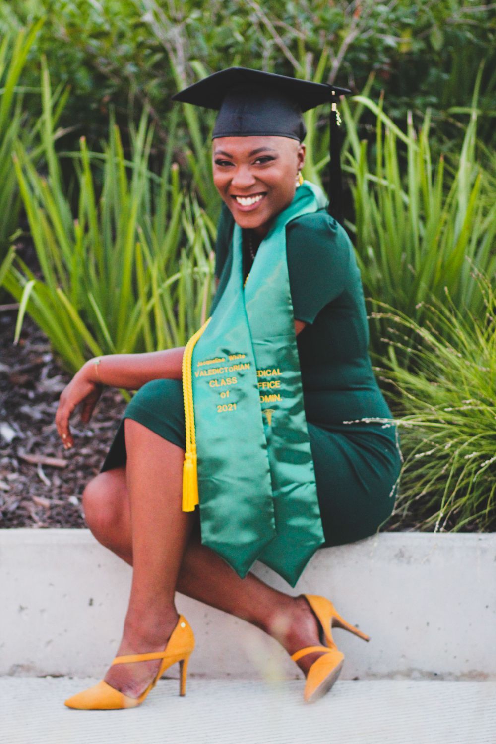 Graduate in emerald green cap and gown with yellow accents poses outdoors.
