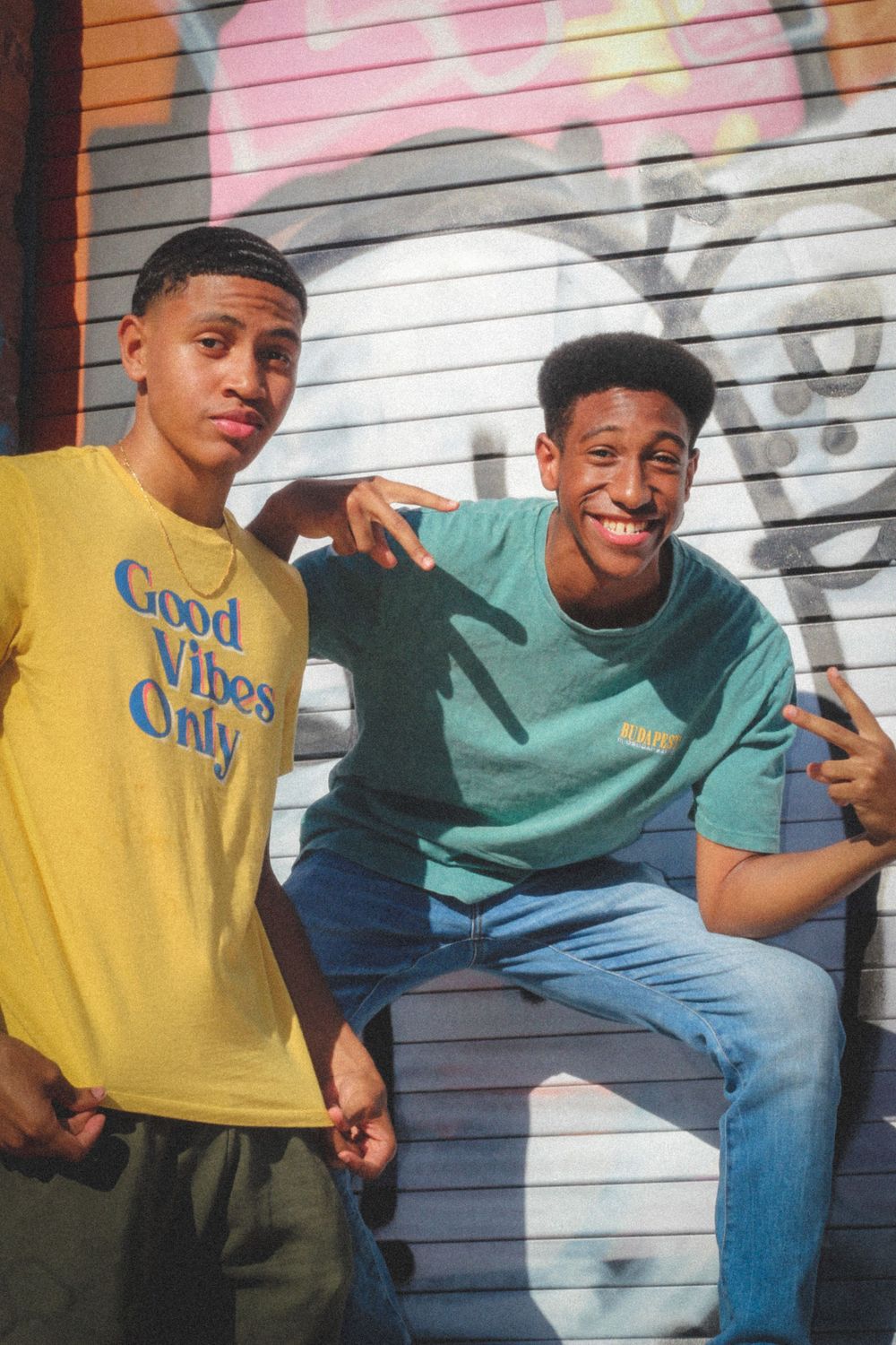 Two friends posing together against graffiti wall wearing colorful t-shirts.