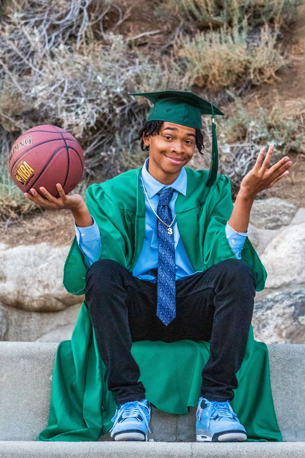 Graduate in green gown holds basketball while sitting on concrete steps outdoors.