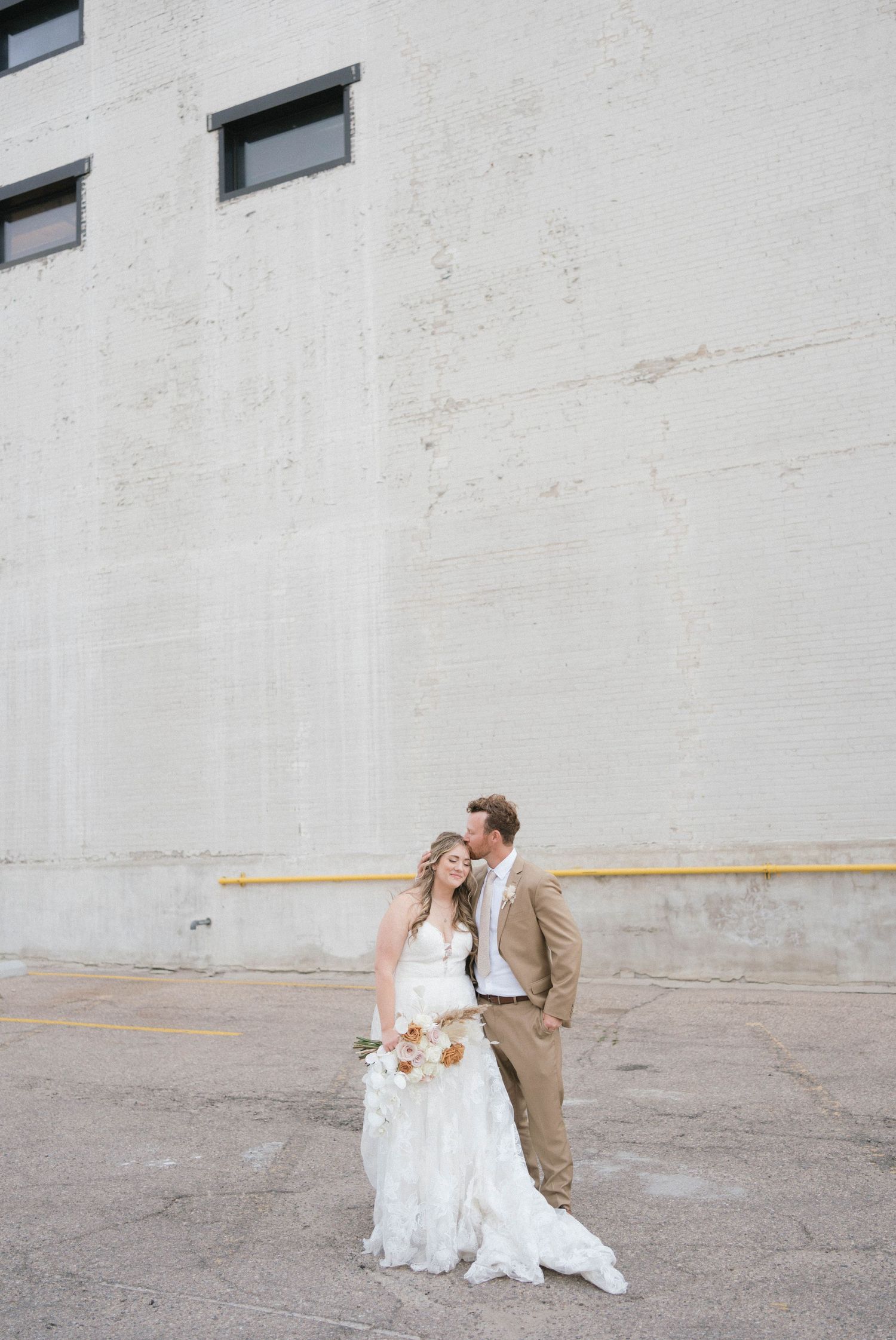 Wedding couple posing against white industrial wall in urban setting.