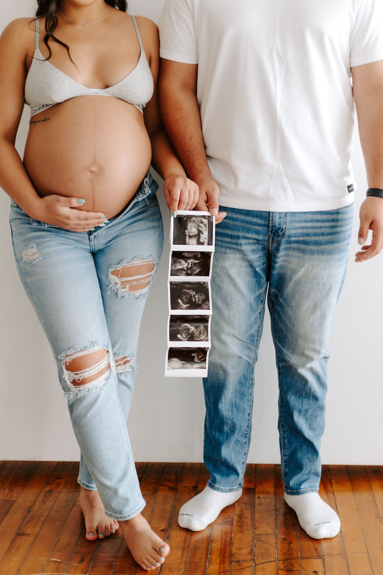 Expectant couple holding ultrasound photos while posing against white wall in matching denim. Calvin Klein maternity shoot.