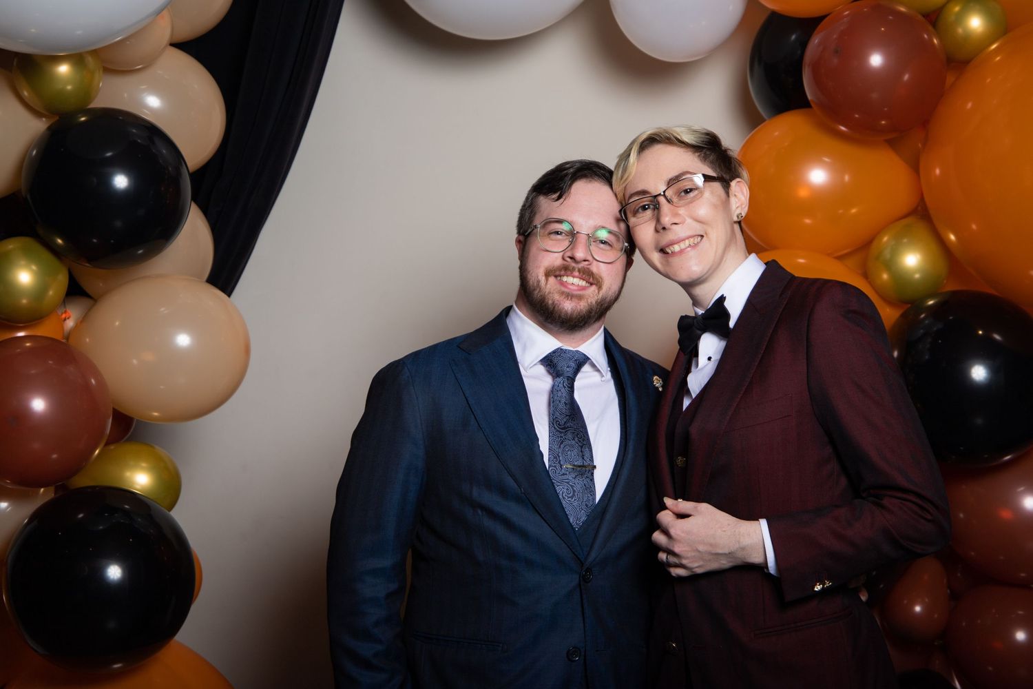 A newlywed couple poses together at a party with orange and black balloon decorations in the background.