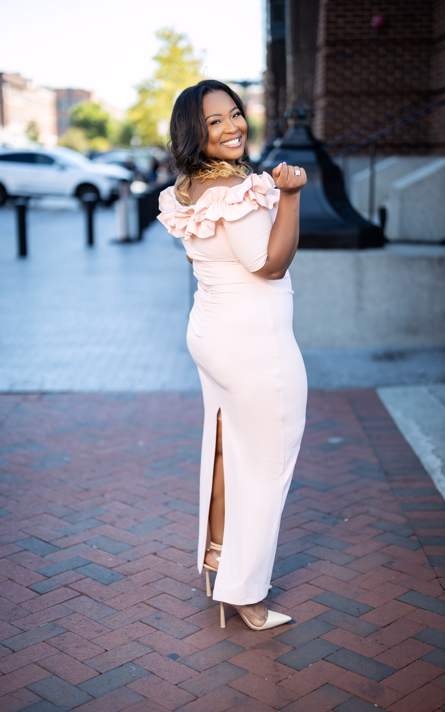 Woman in pink dress with side slit poses playfully on brick pavement.