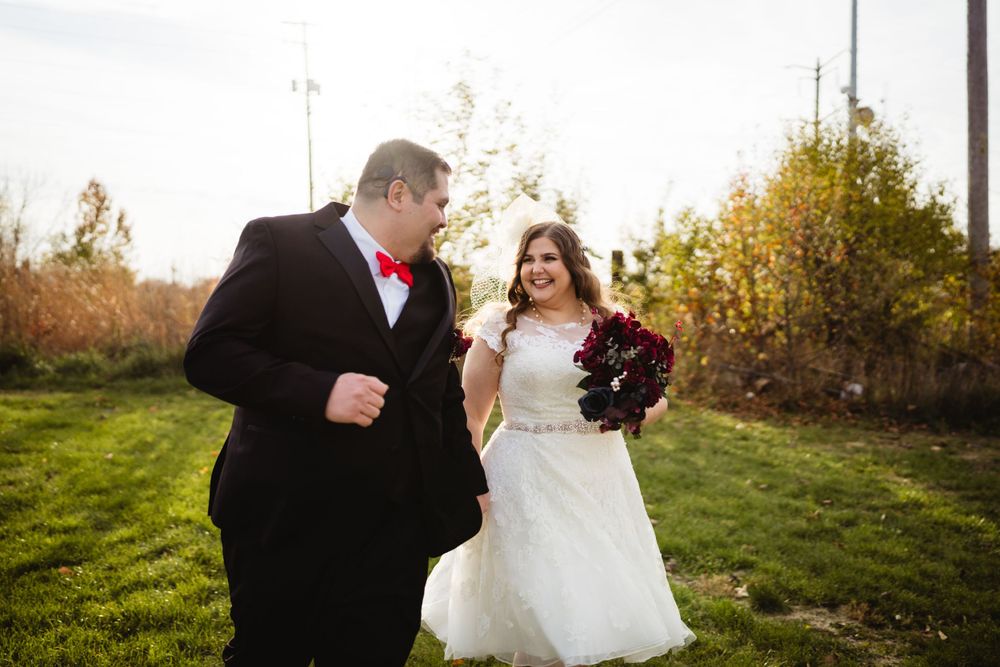 A couple in wedding attire shares a joyful moment together outdoors on a fall day with autumn foliage in the background.