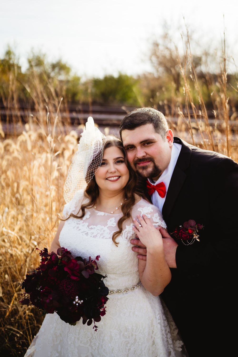 Wedding couple posing together in field with burgundy floral accents.