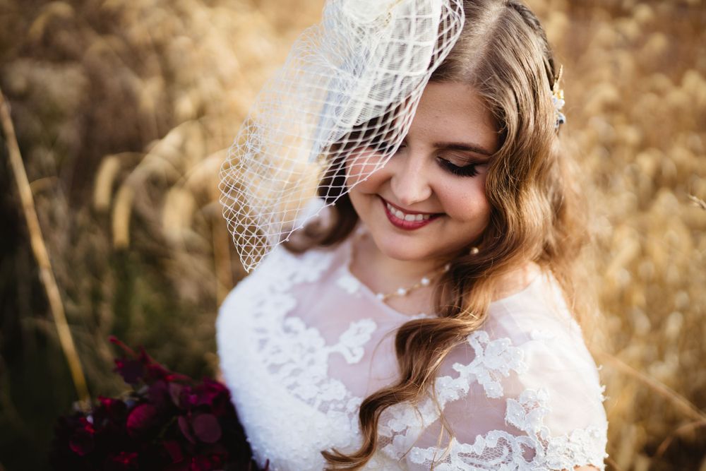 Bride in lace dress and birdcage veil poses in golden field.