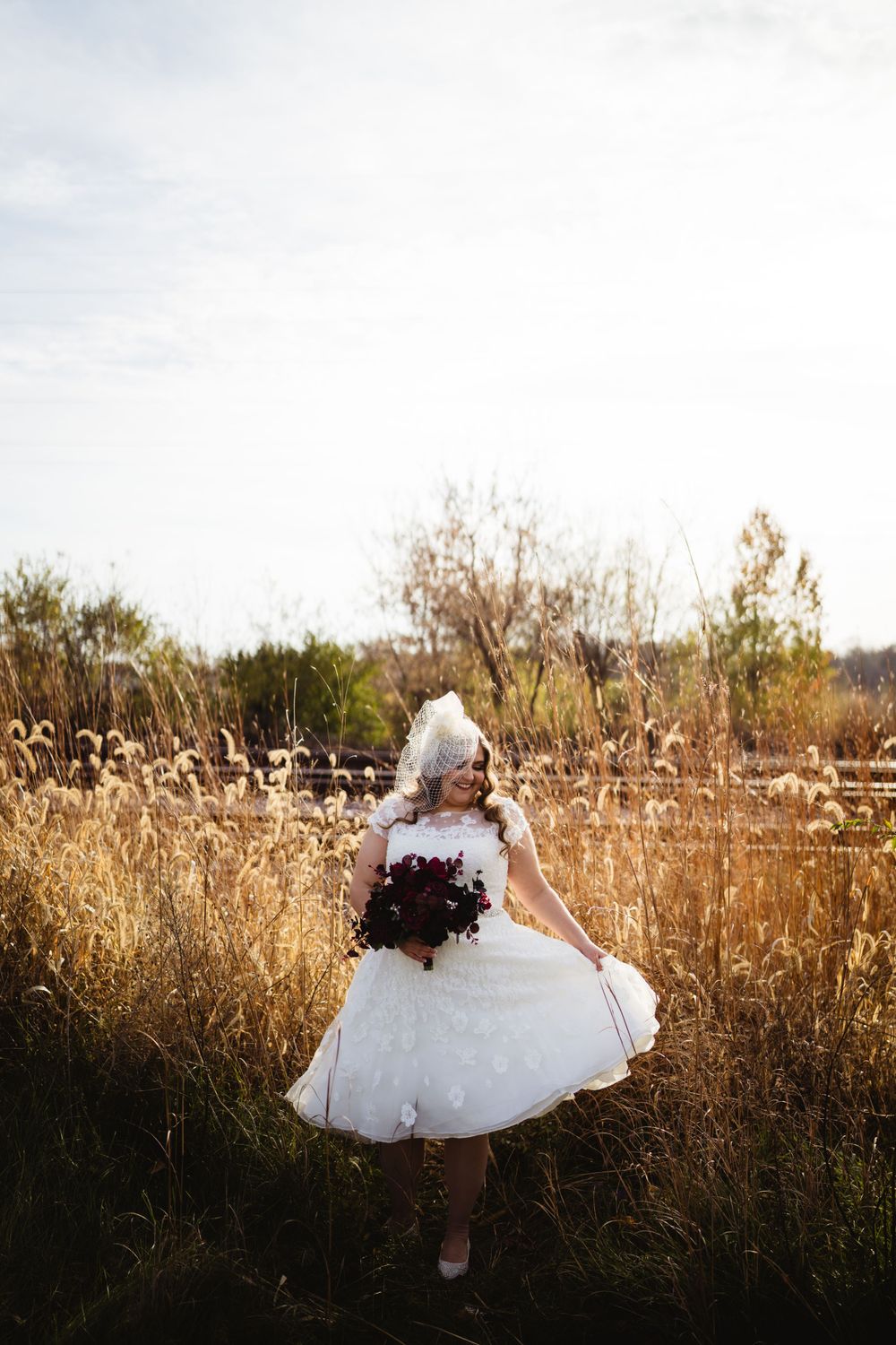 Bride in short wedding dress holding burgundy bouquet in sunlit field.