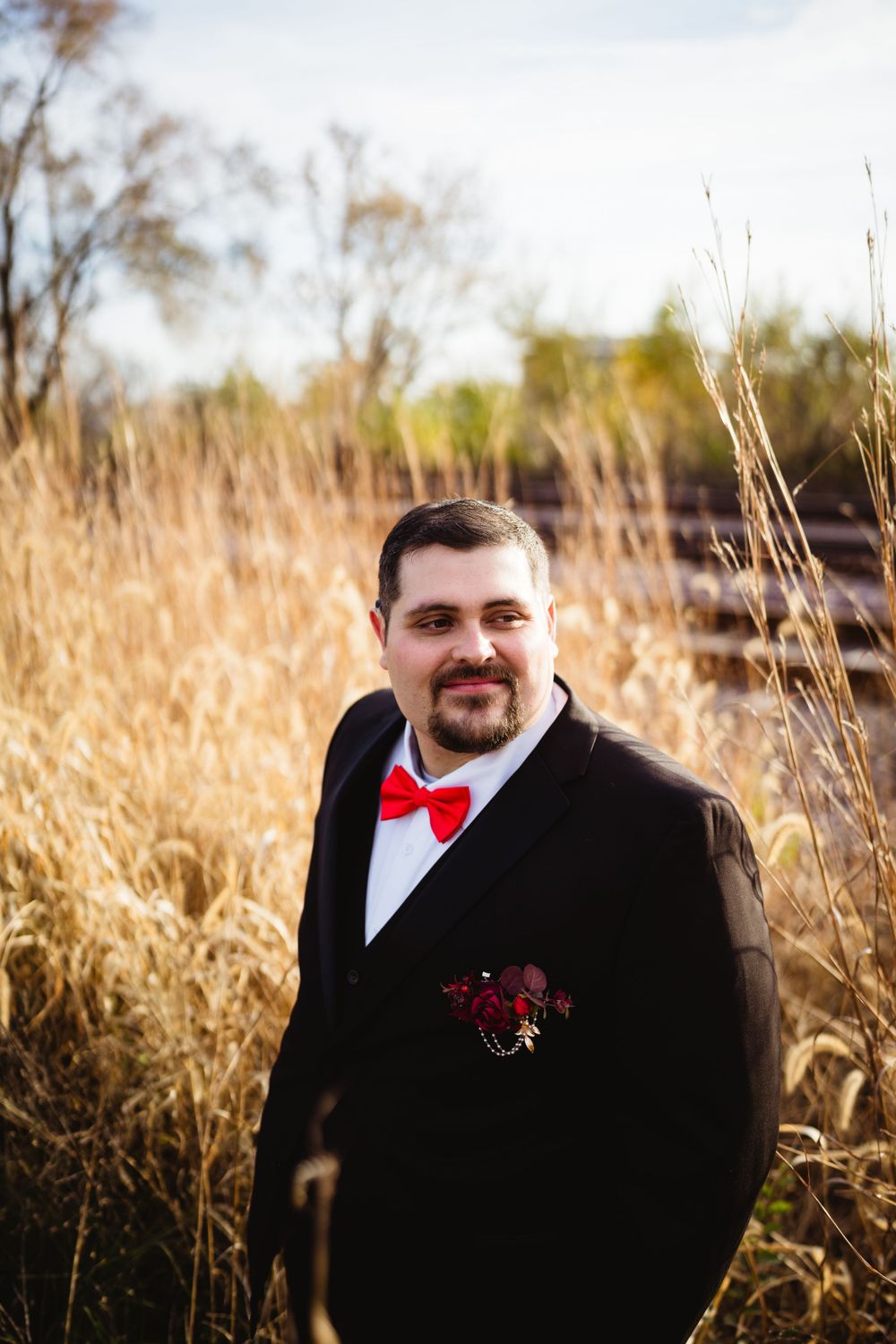 Groom in black tuxedo and red bow tie poses in autumn field.