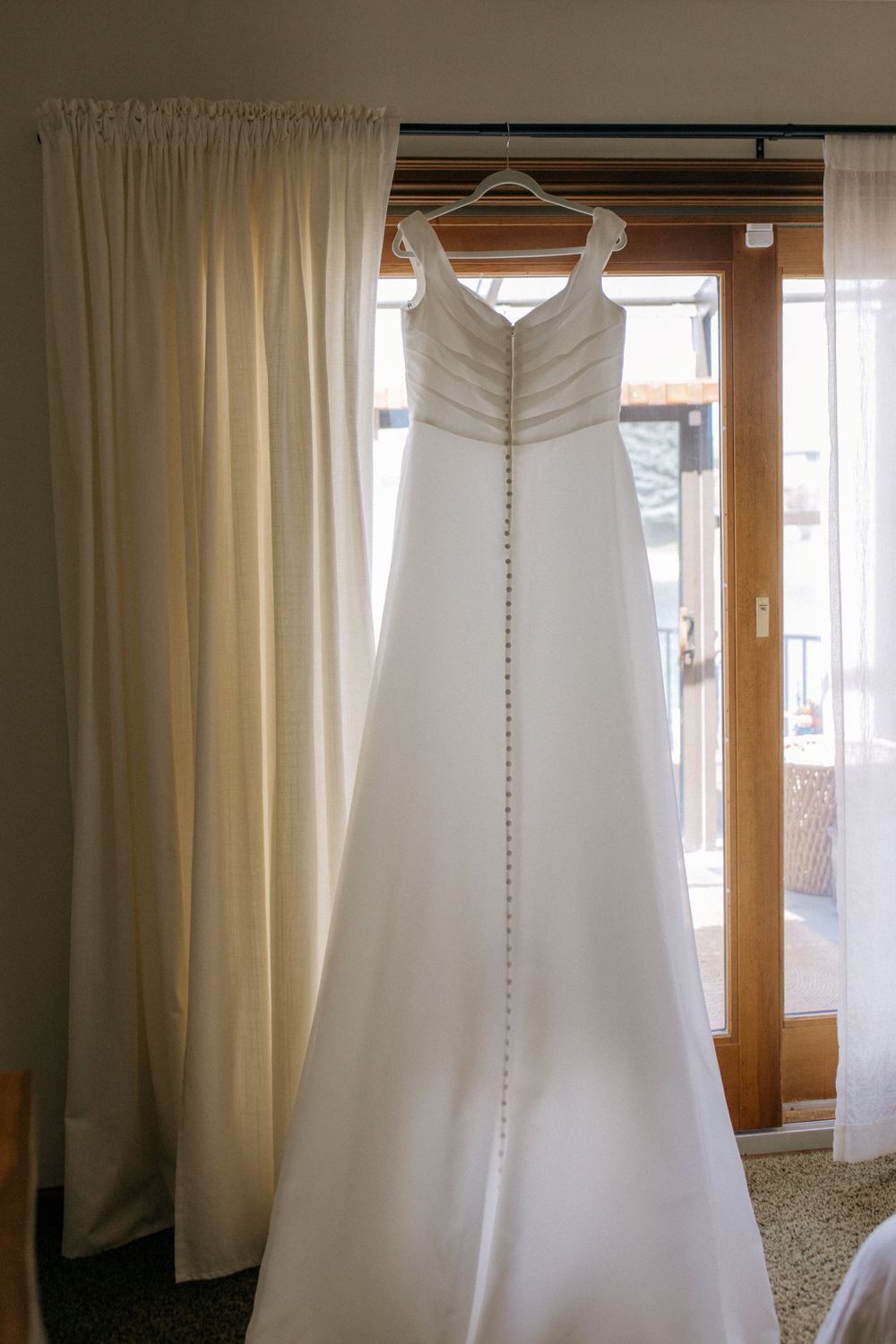 Classic white bridal gown with button detail down back hanging against cream colored curtains.
