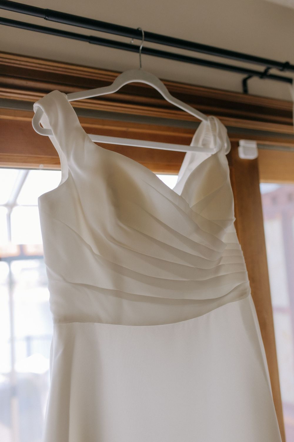Close-up detail of pleated bodice on white wedding gown hanging by window.