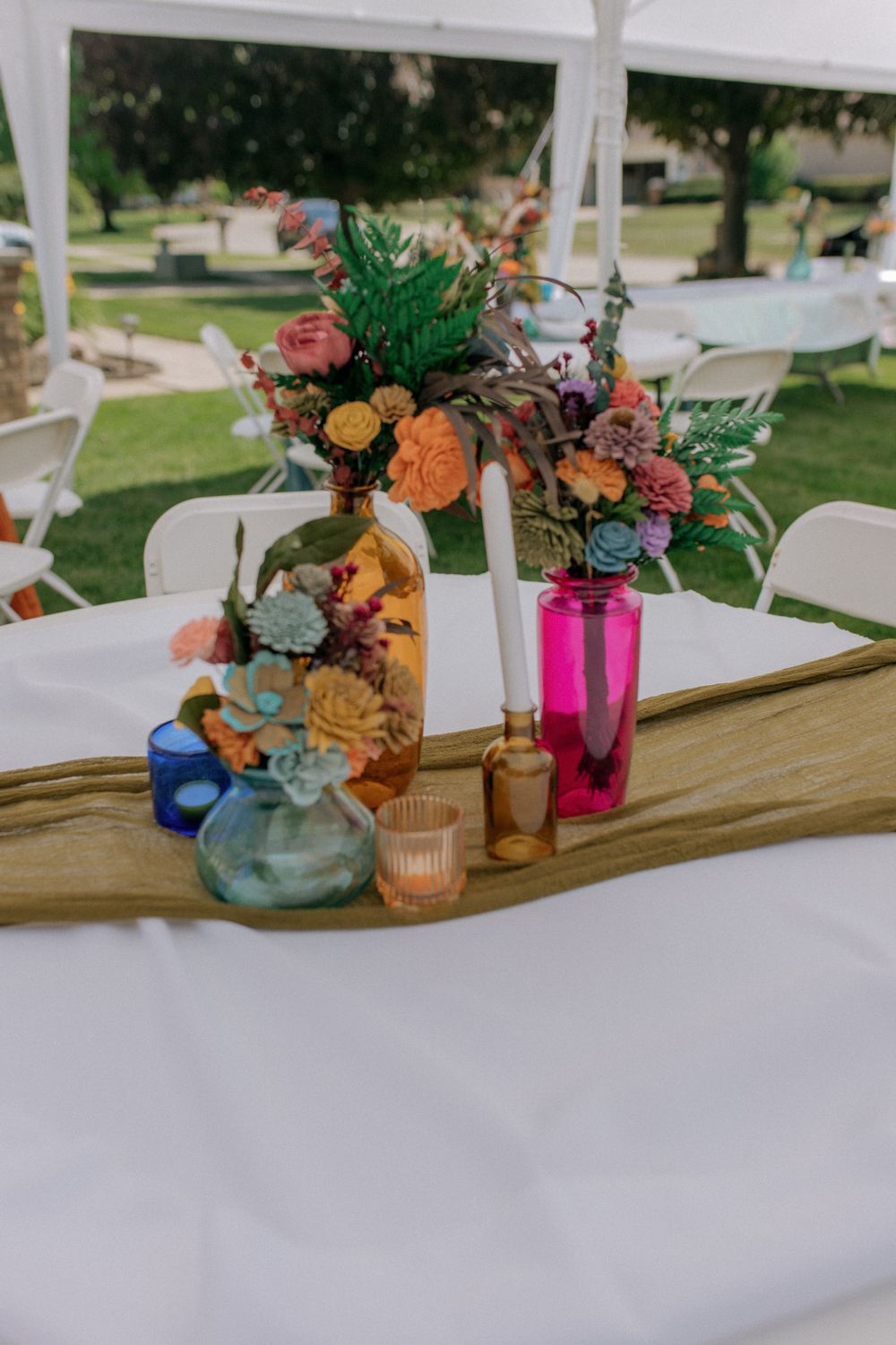 Wedding reception table decor featuring colorful flowers in mixed glass vases on wooden plank.