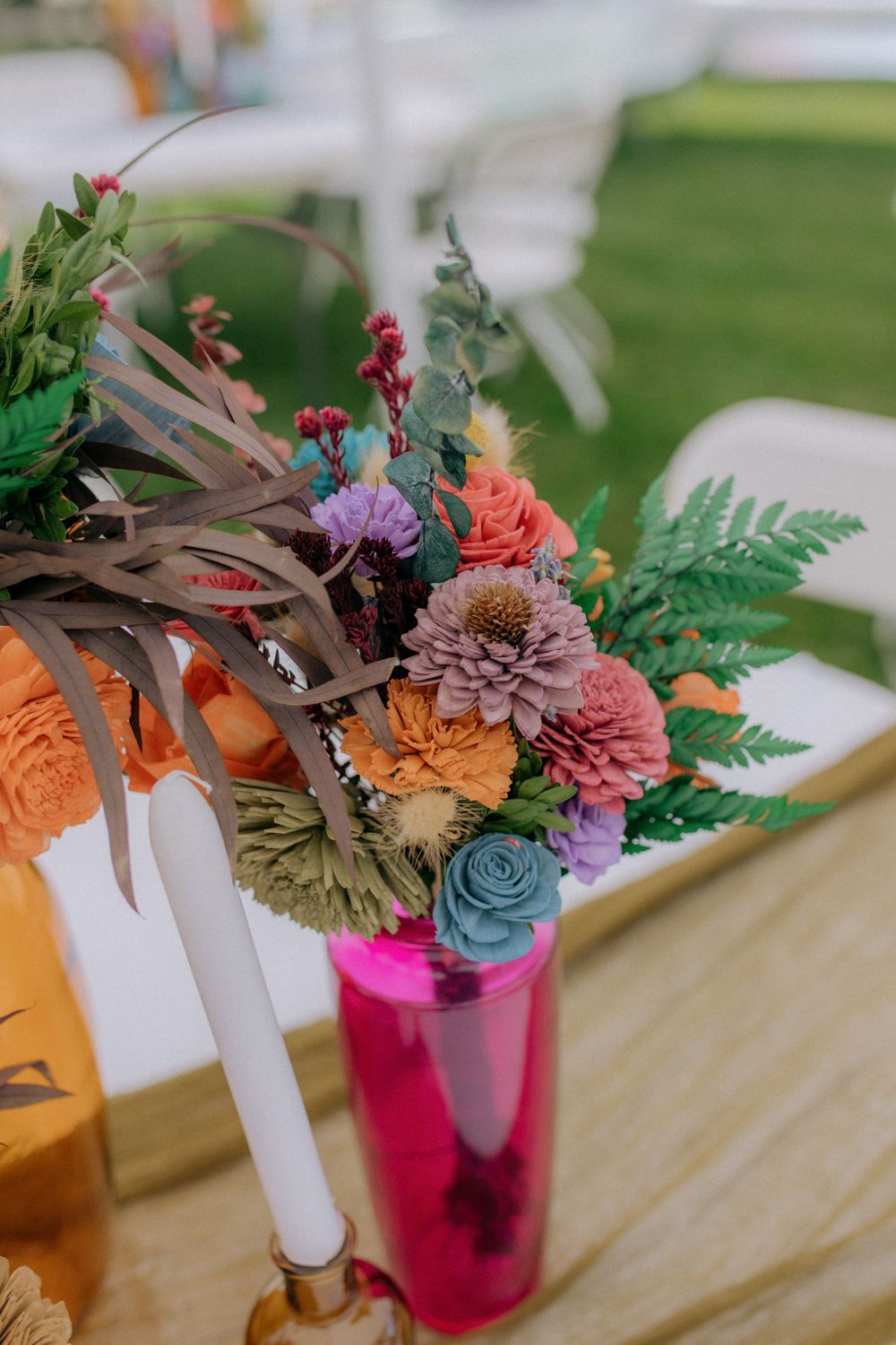 Colorful floral arrangements in various bottles sit atop a wooden table at an outdoor event.