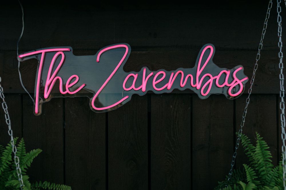 Pink neon sign reading 'The Zarembas' mounted on dark wooden wall with decorative hanging plants.