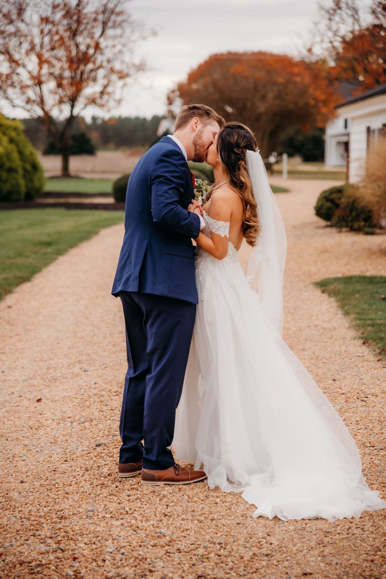 Romantic wedding couple shares intimate moment on gravel path surrounded by autumn trees.