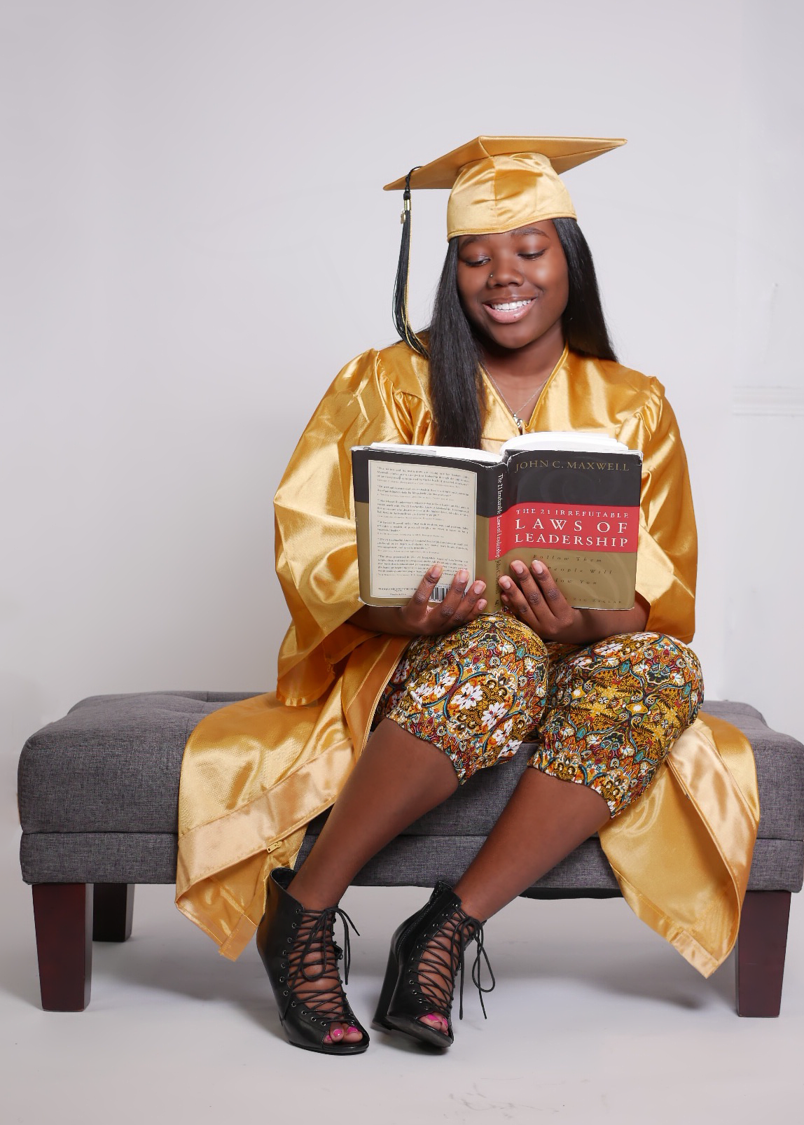 Graduate in gold regalia reads books while seated on grey bench.