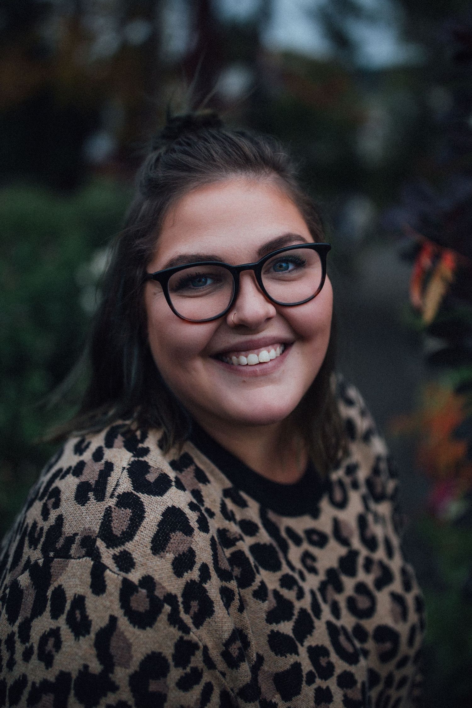 a close portrait of a femme with glasses, a nose ring, large smile and leopard print sweater.