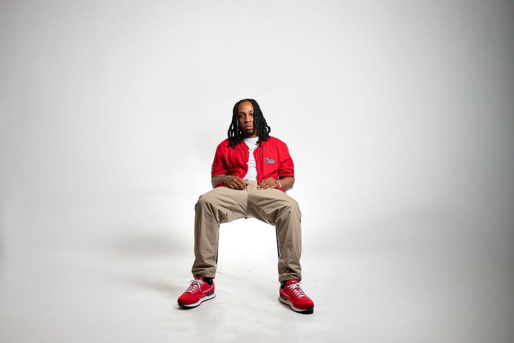 Person in red jacket and khaki pants sitting on chair against white background wearing red sneakers.