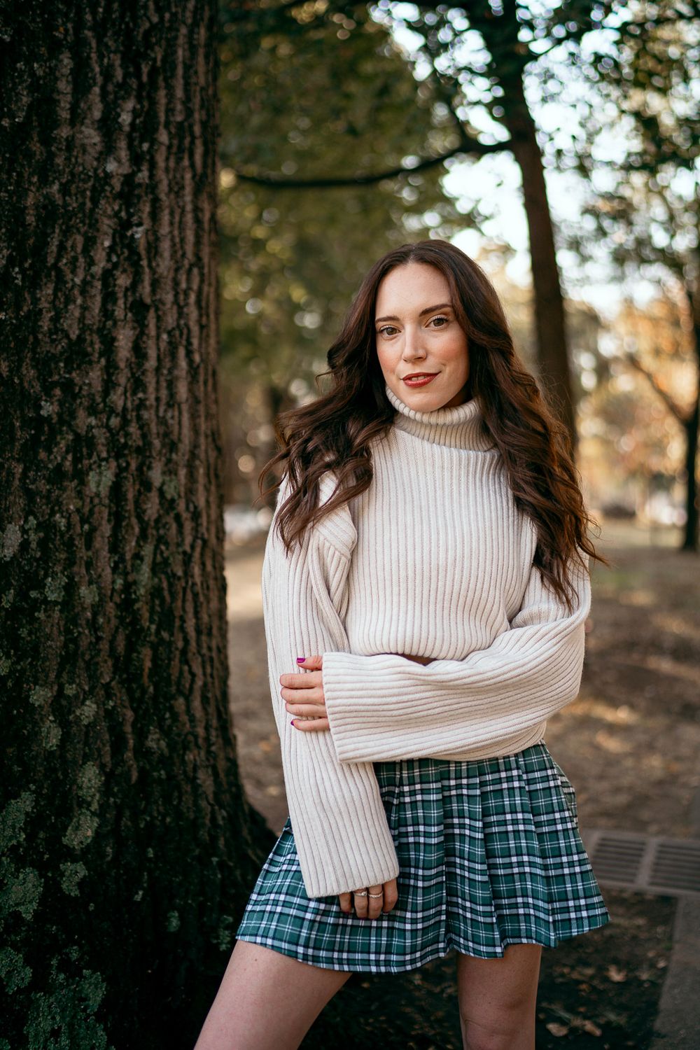 A cozy fall fashion look featuring a cream turtleneck sweater and plaid skirt photographed in a forest setting.