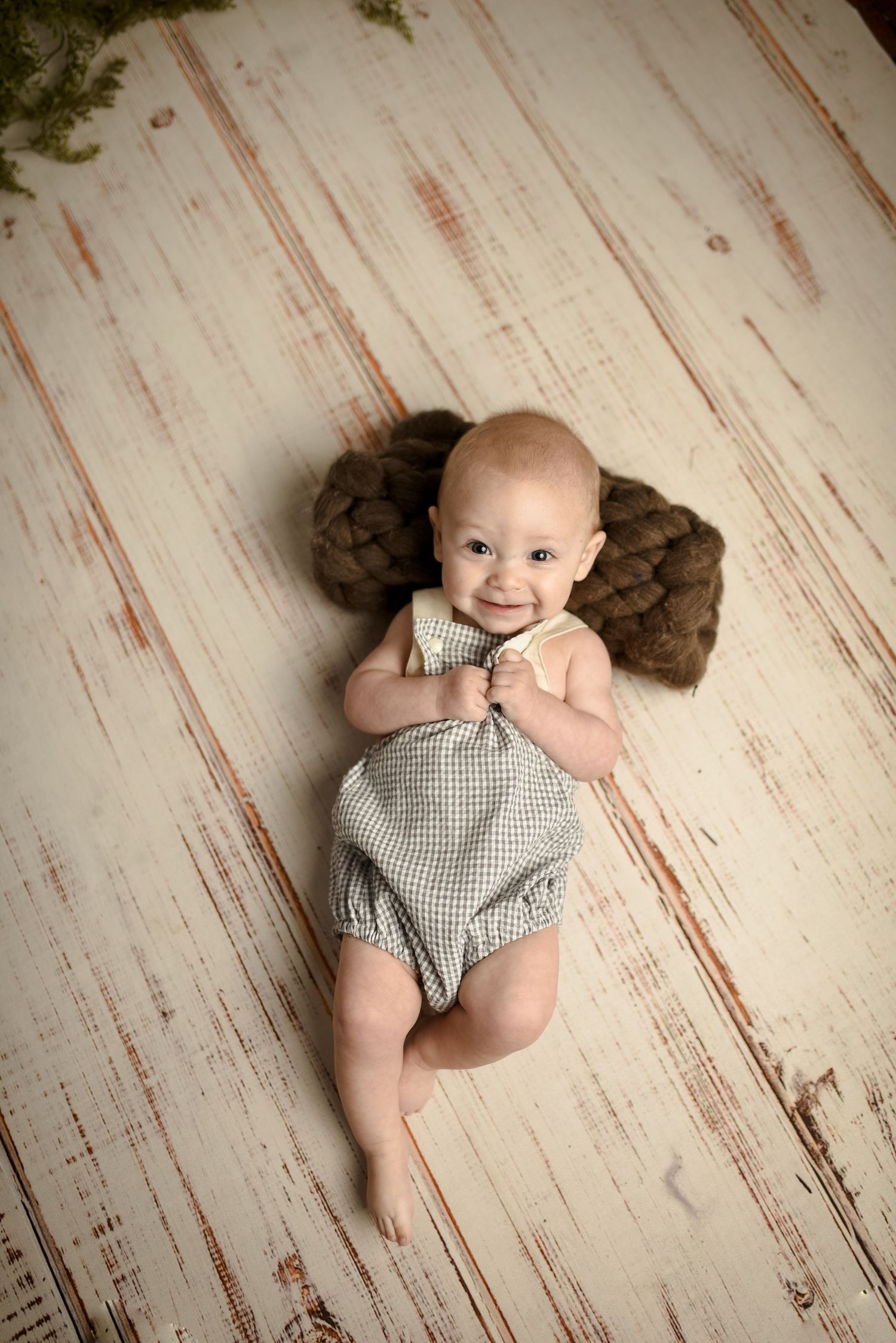 Baby in gray gingham romper lying on distressed white wooden floor.