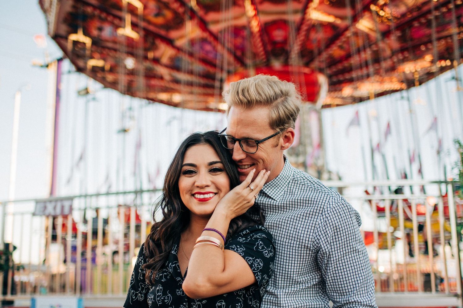 A happy couple shares an intimate moment in front of a colorful carousel at an amusement park.