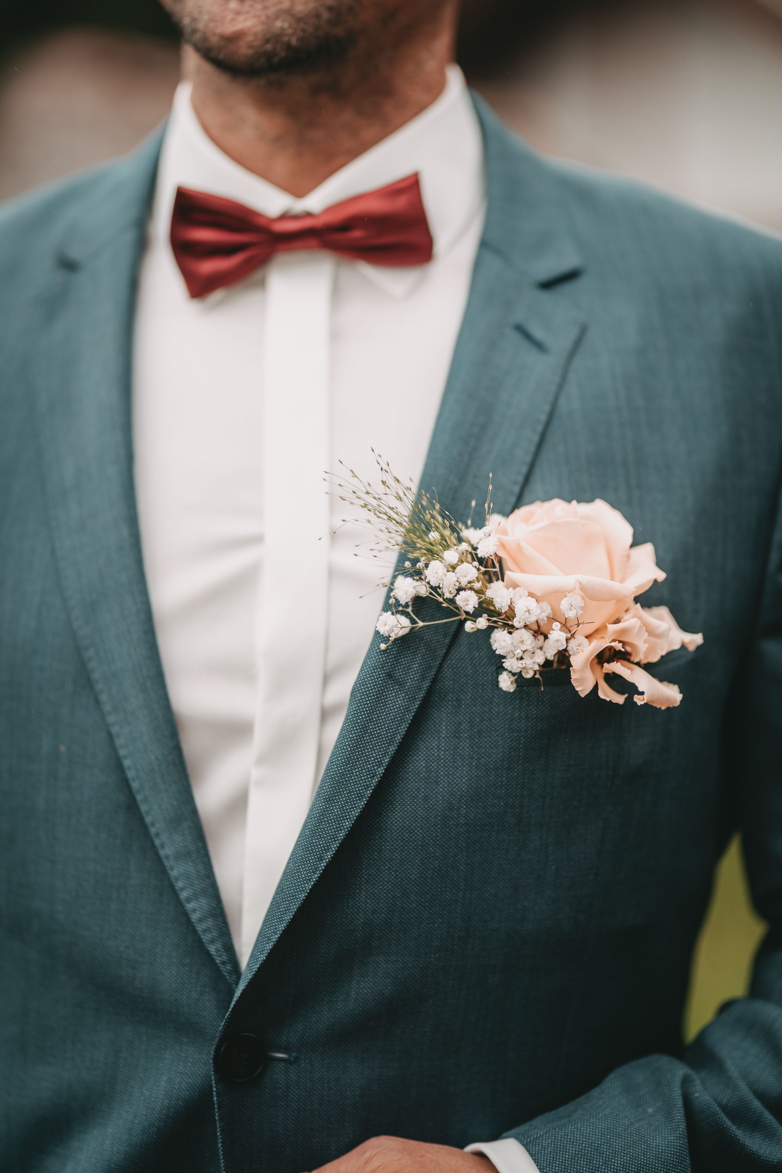 Close-up of a groom's boutonniere with peach roses and greenery against a teal suit jacket and burgundy bow tie.