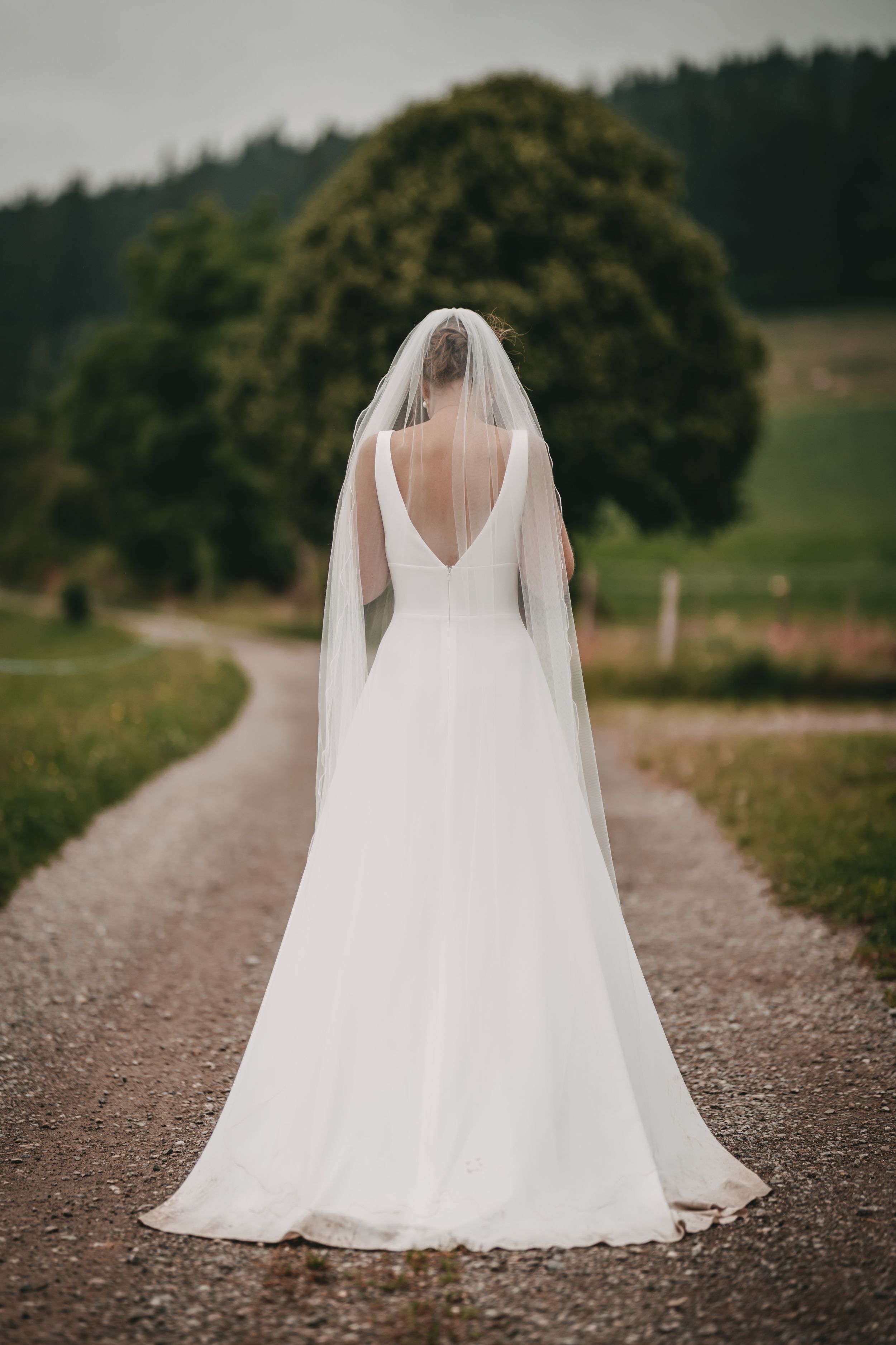 Back view of a flowing white wedding dress and veil on a gravel path with mountain scenery.