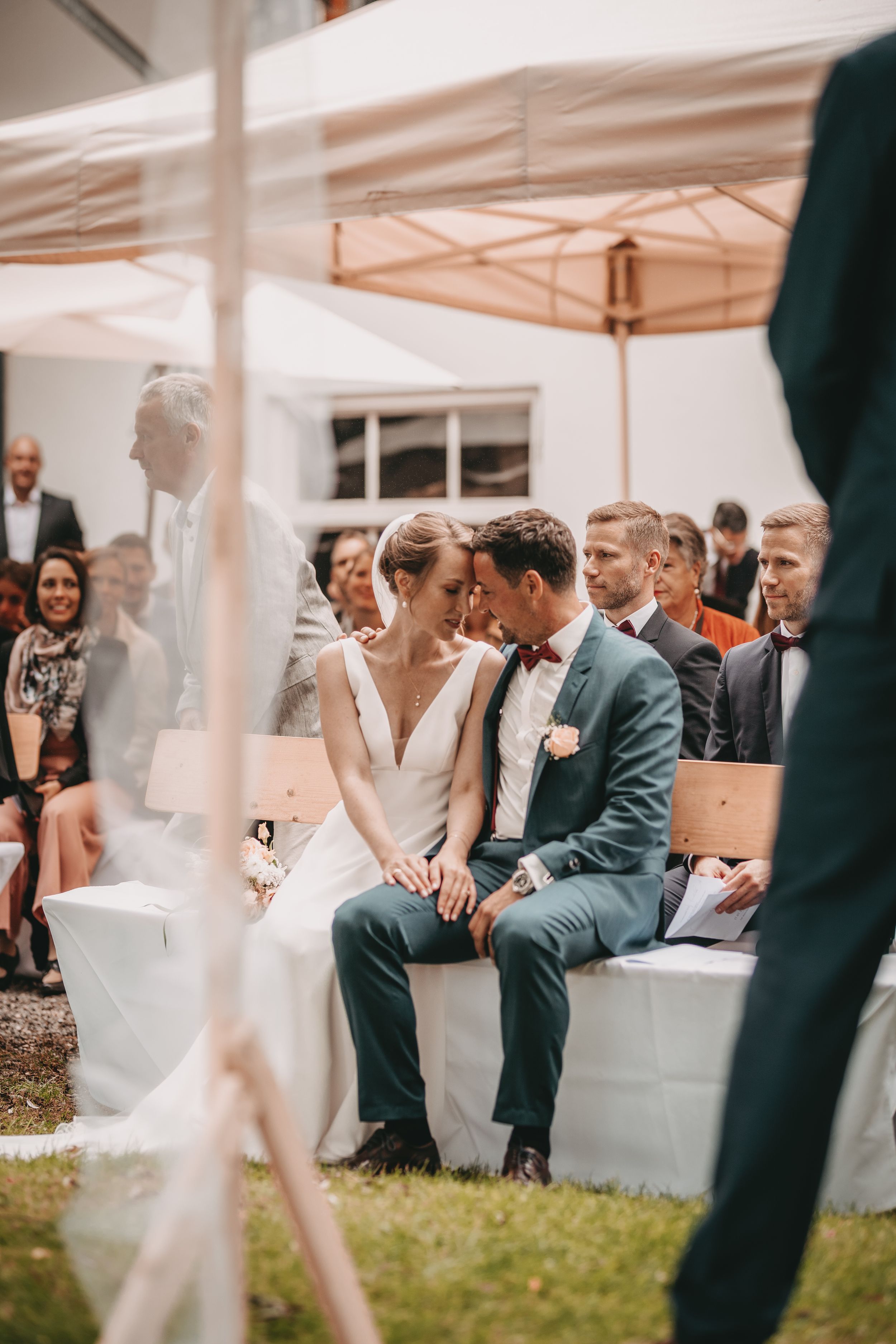 Outdoor wedding ceremony under white tents with guests seated on white chairs.