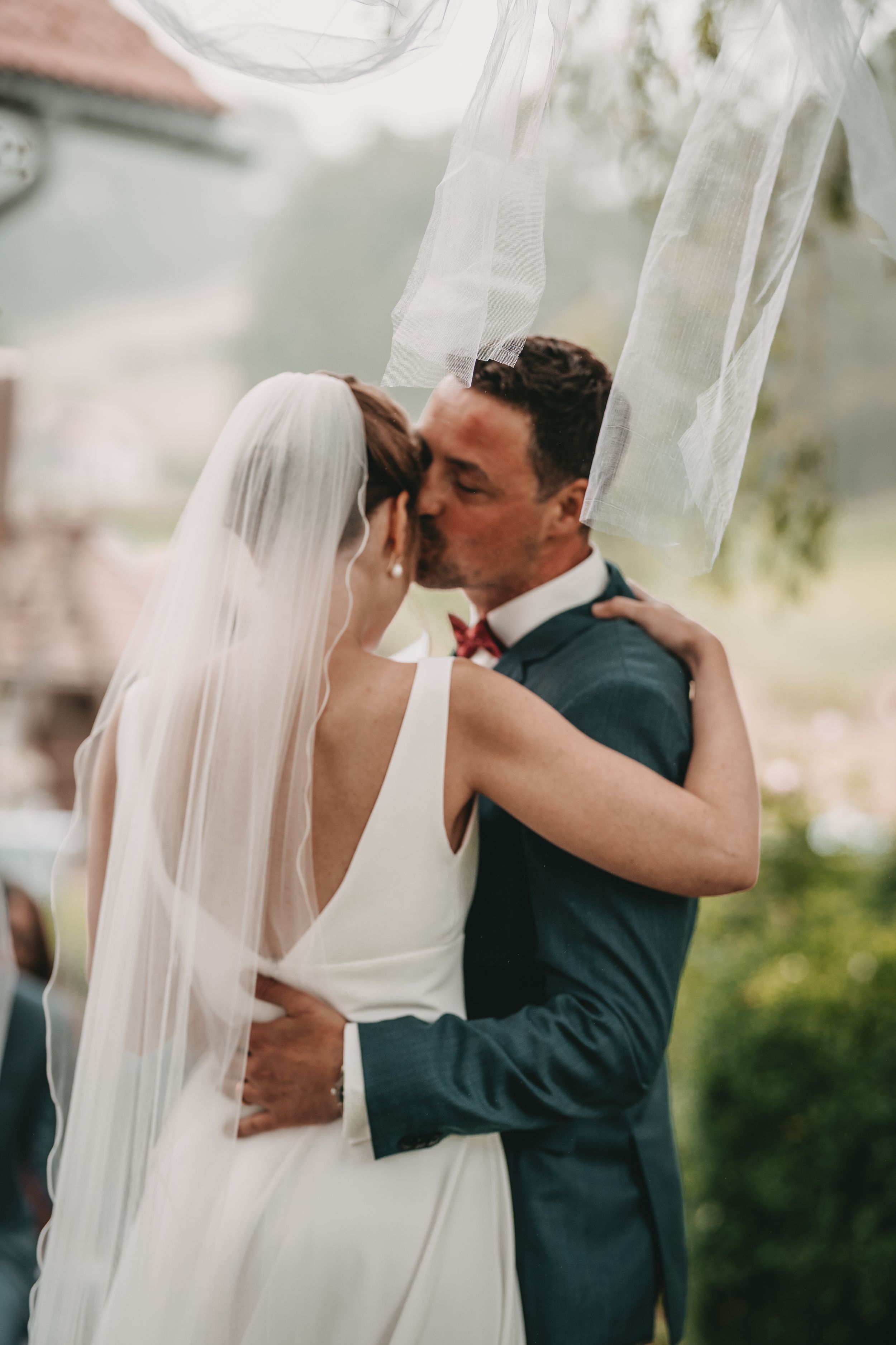 Newlywed couple shares a romantic kiss under a flowing veil during their outdoor wedding ceremony.