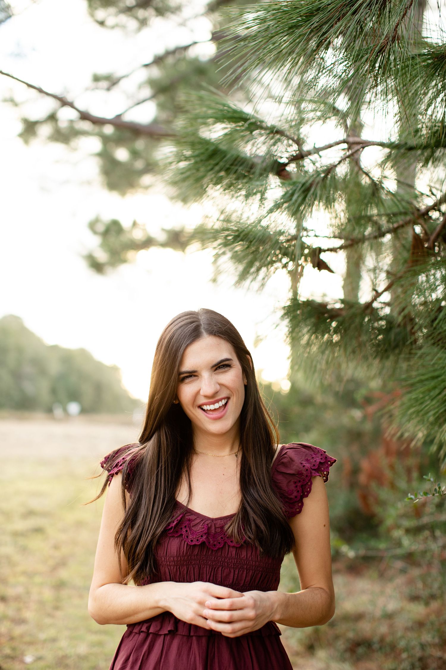 Woman in burgundy dress smiles brightly against a backdrop of pine trees.