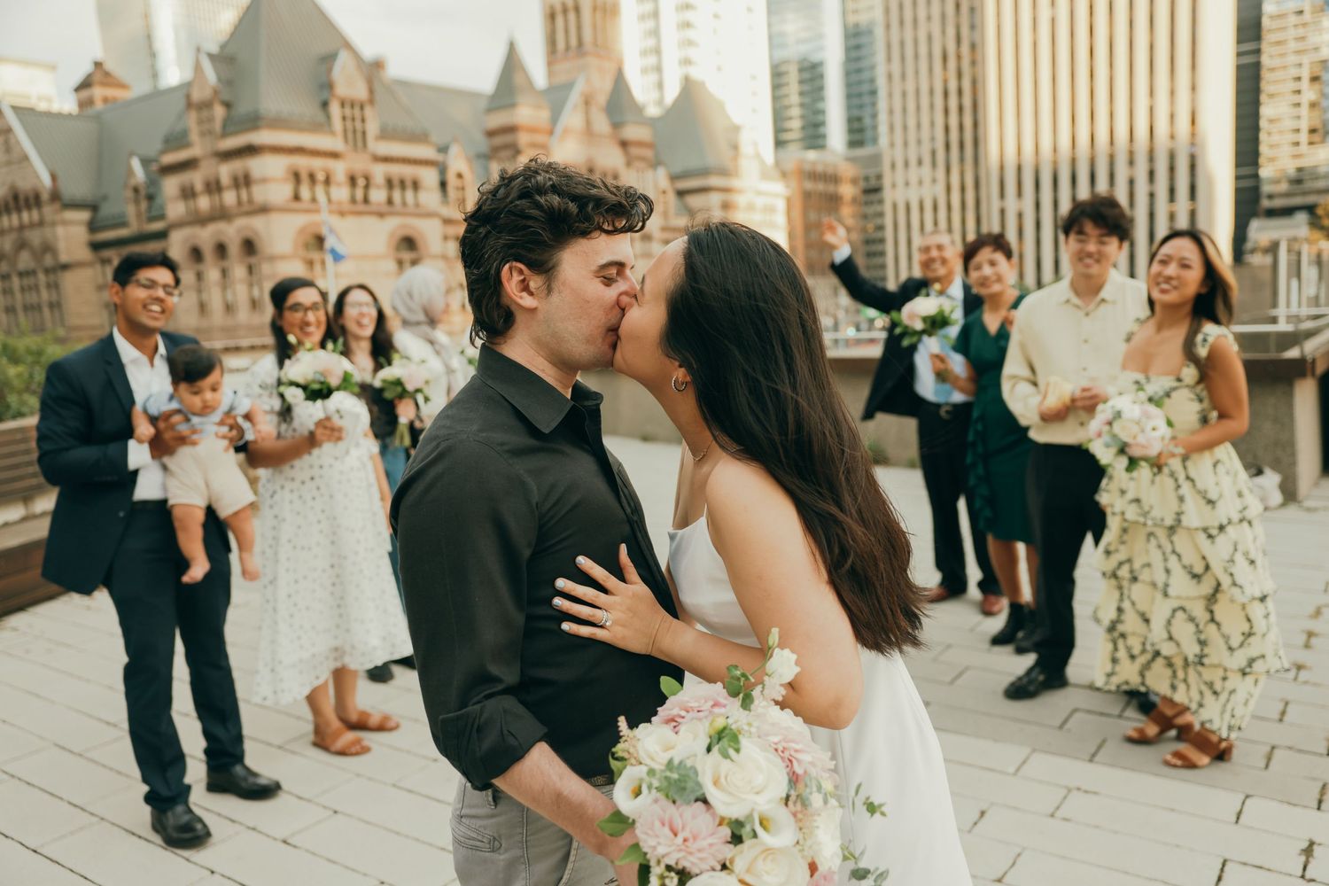 A newlywed couple shares a kiss on a rooftop terrace while wedding guests look on and smile in the background.