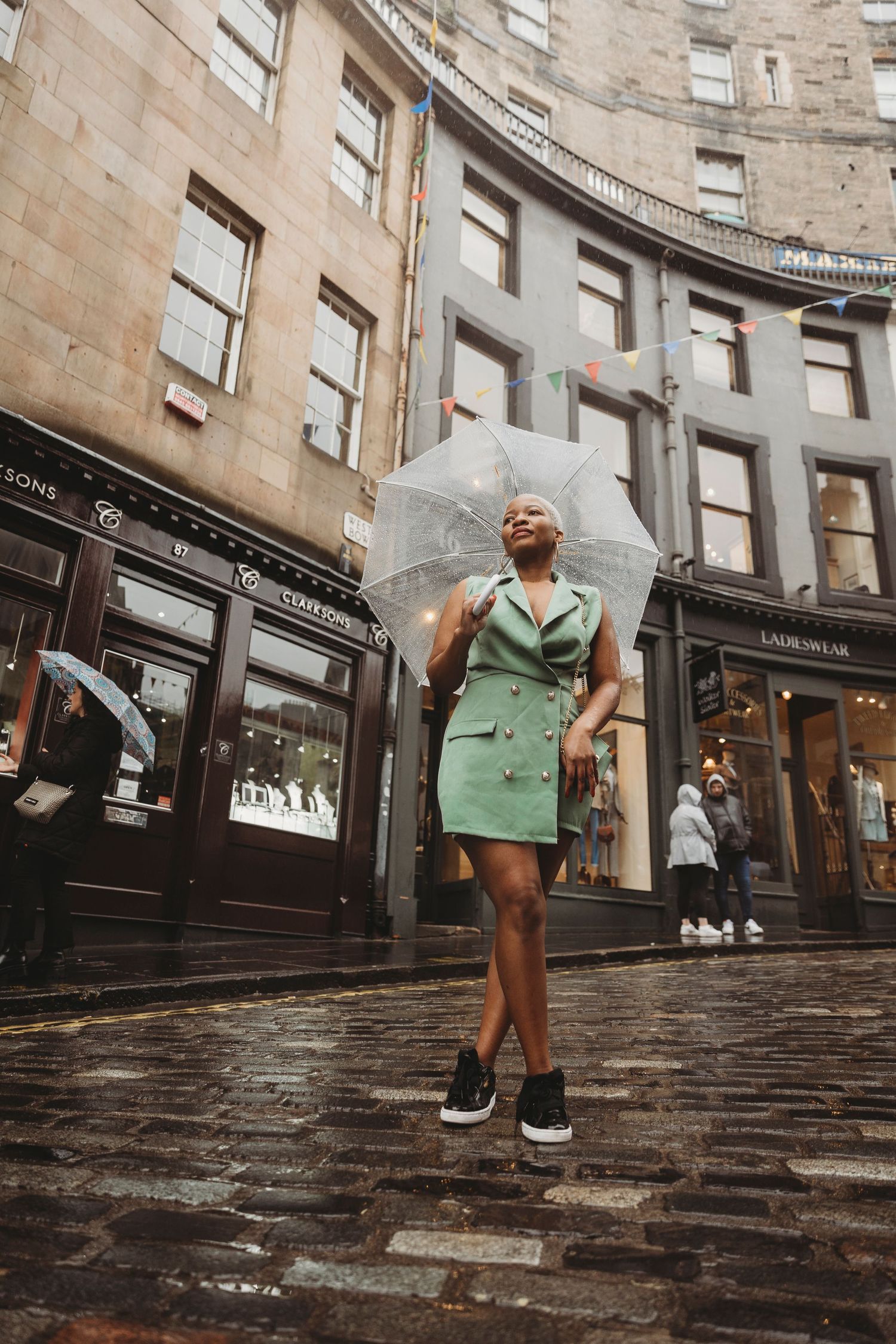 A stylish person holding a clear umbrella poses on wet cobblestone street in front of curved stone buildings.