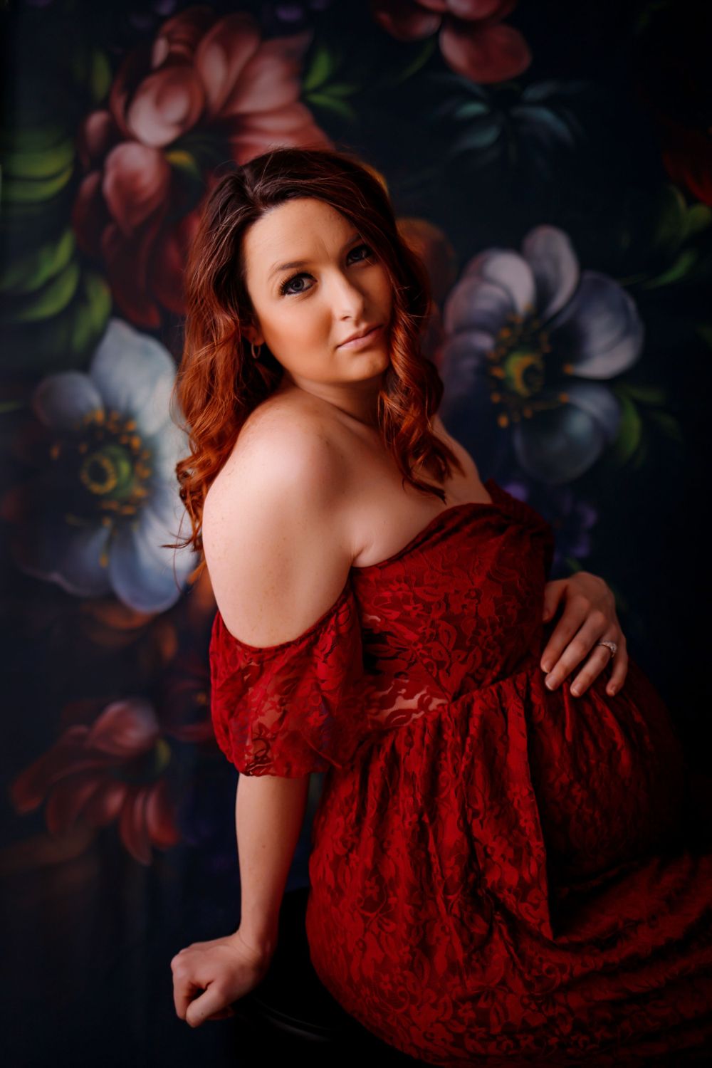 A studio portrait against a dark floral background wearing a red lace dress.