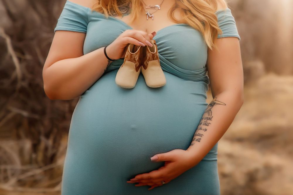 A close-up shot showing tiny baby shoes held against a pregnant belly in a blue dress.
