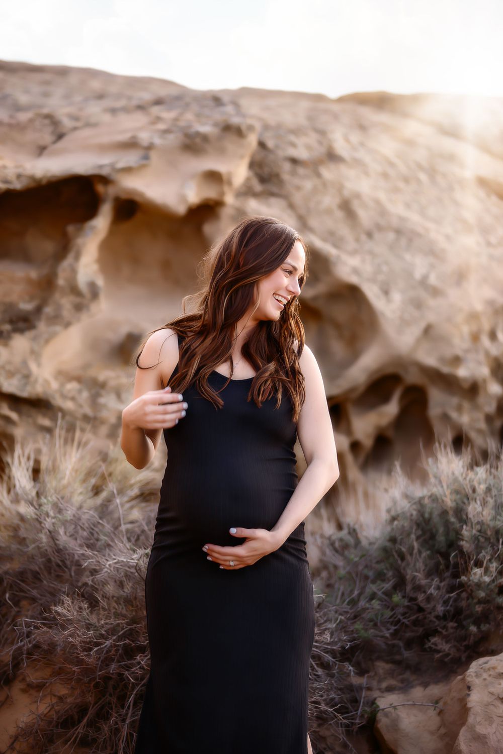 A maternity portrait in a black dress against desert rock formations at sunset.