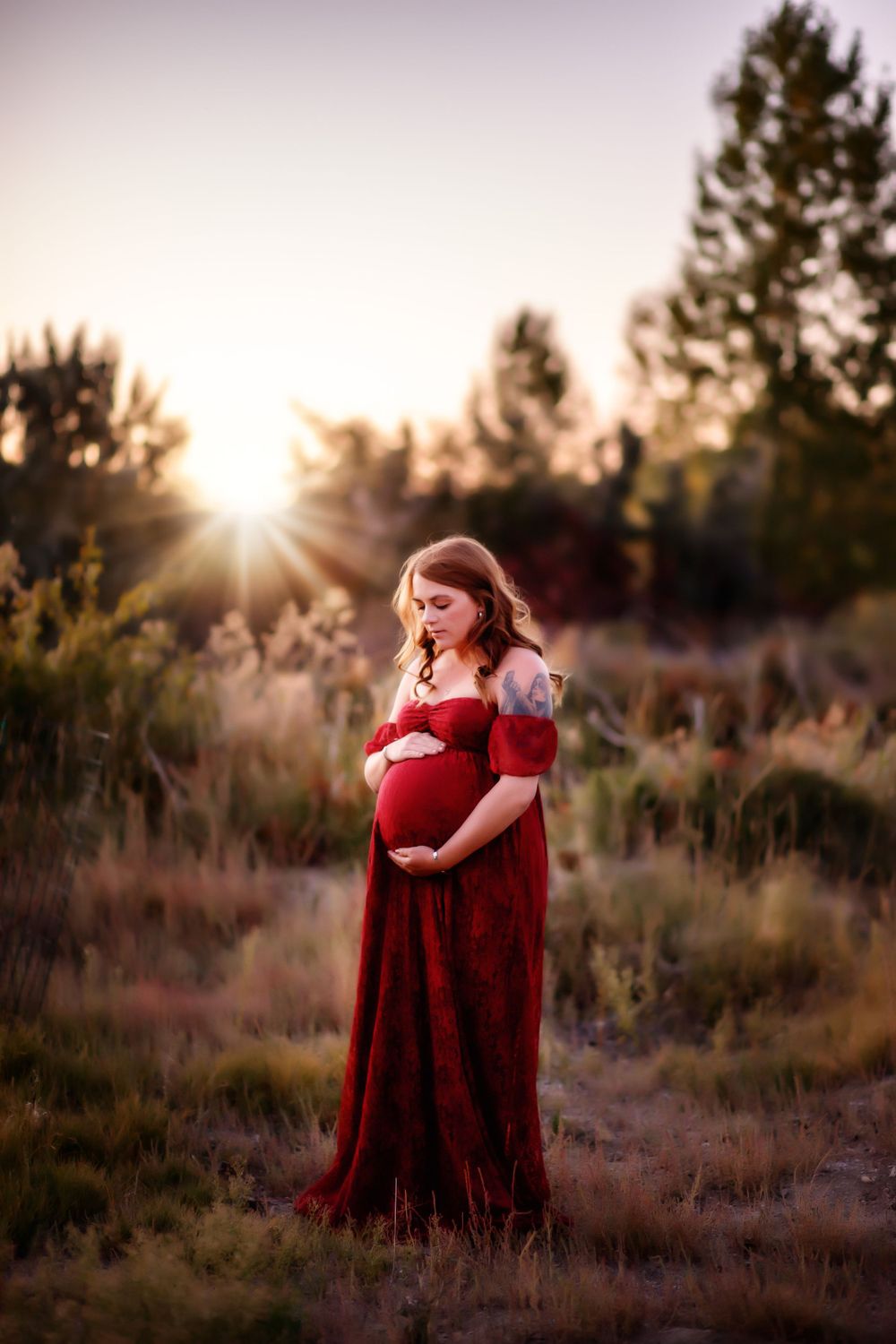 A maternity photo in a red dress against a sunset backdrop with tall grass and pine trees.