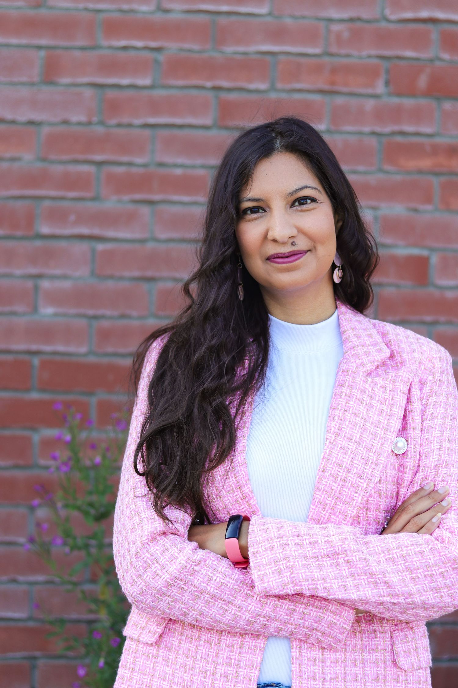 Confident pose in pink tweed jacket against rustic brick wall with statement earrings.