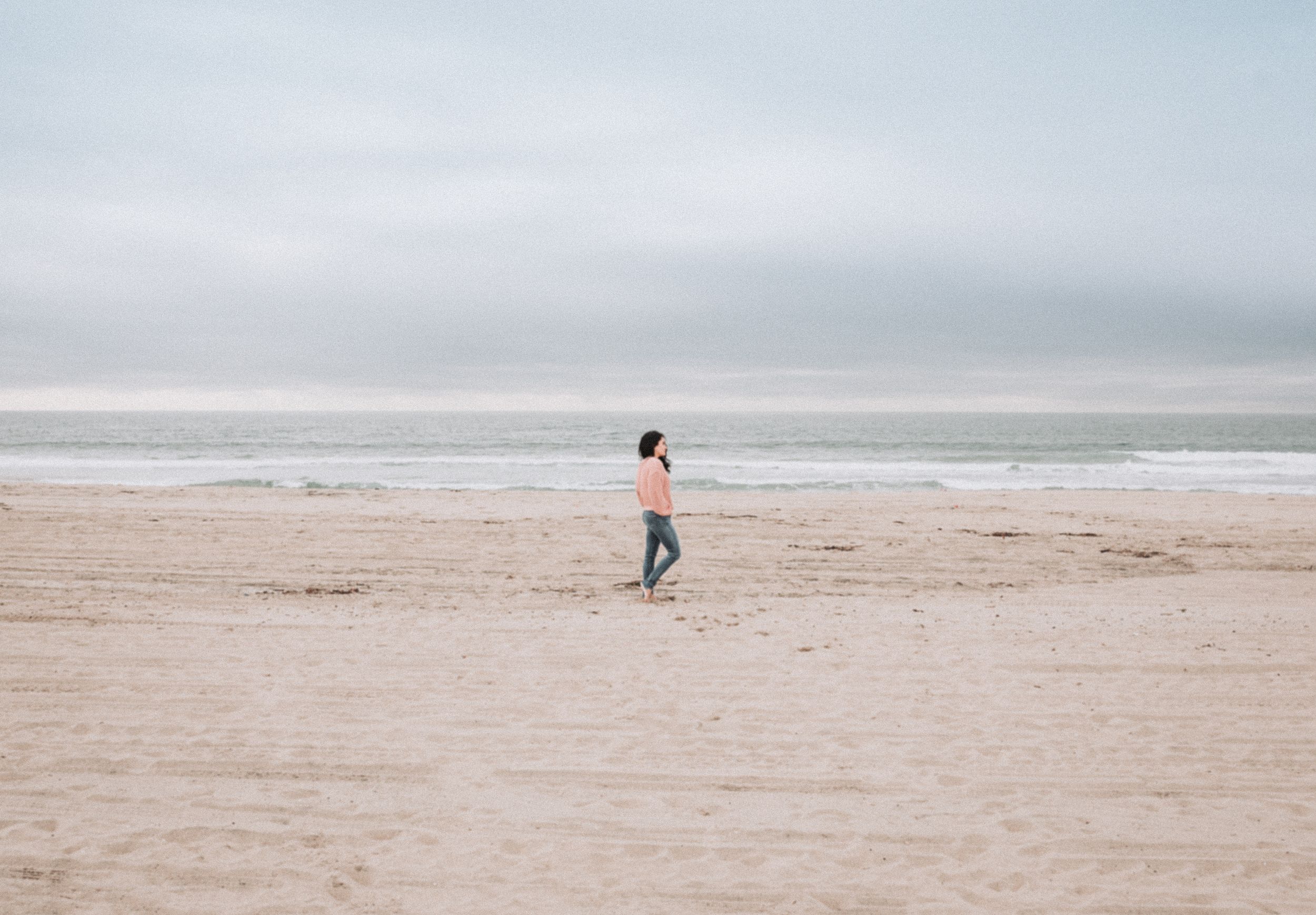 A lone figure walks along a misty beach shore on an overcast day.