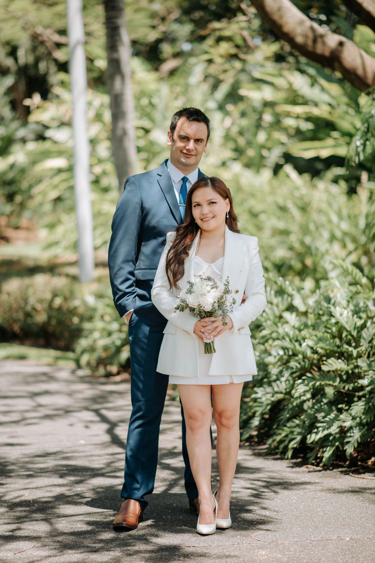 Stylish couple poses on garden path with bride in modern white blazer dress and groom in blue suit.