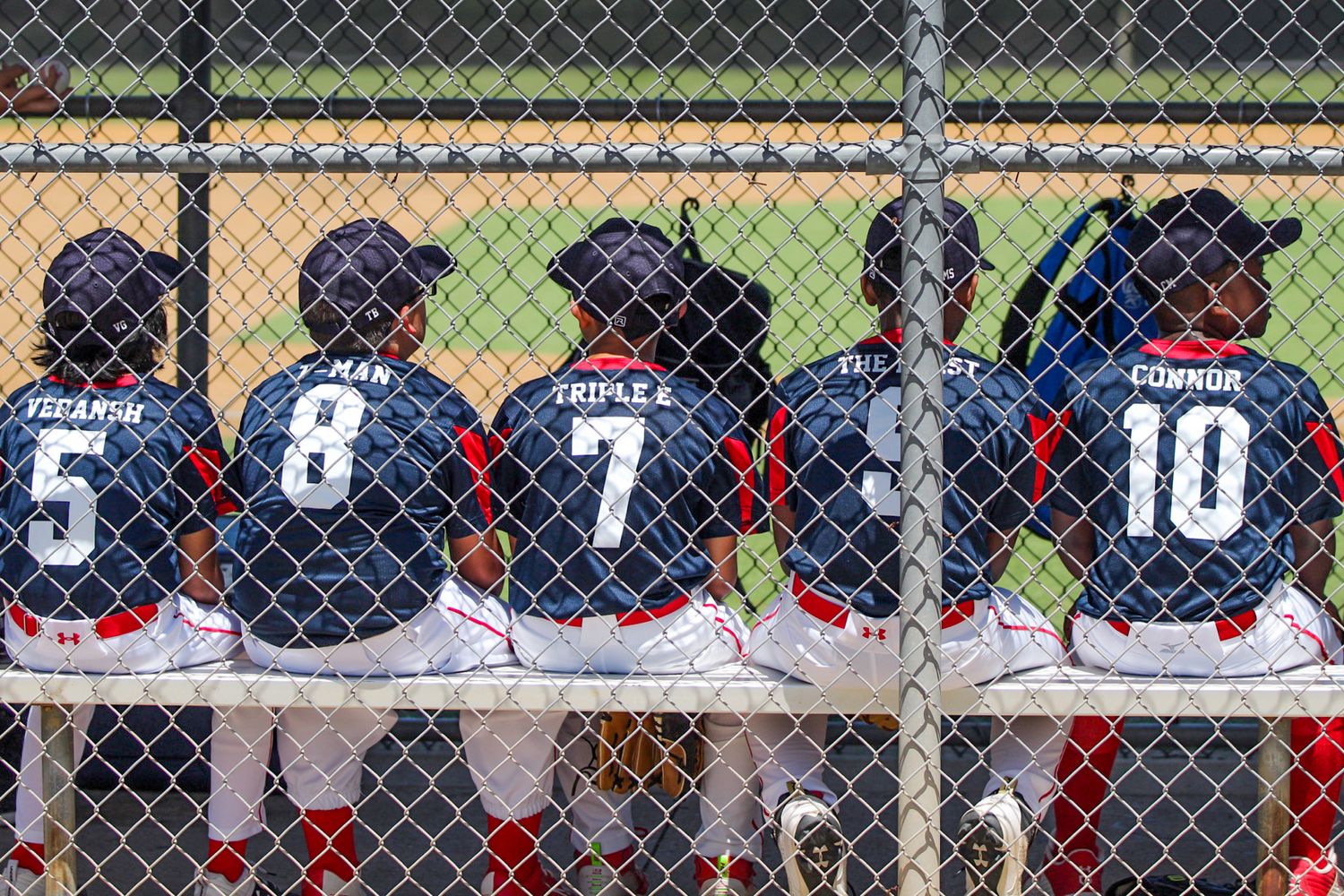 Baseball players in navy uniforms with jersey numbers 5, 8, 7 and 10 watching game from behind chain link fence.
