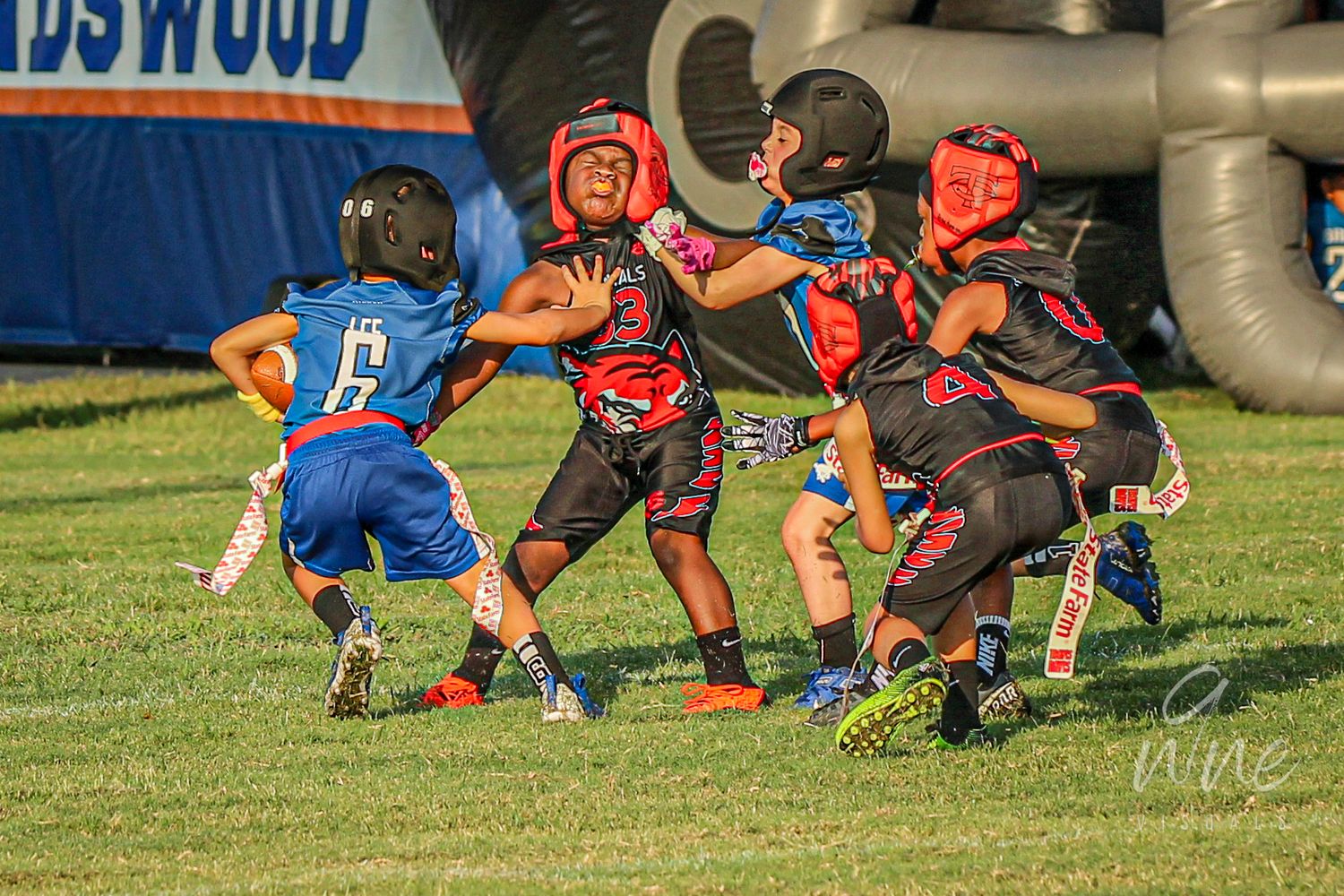 Youth flag football players in protective gear competing on sunny grass field.