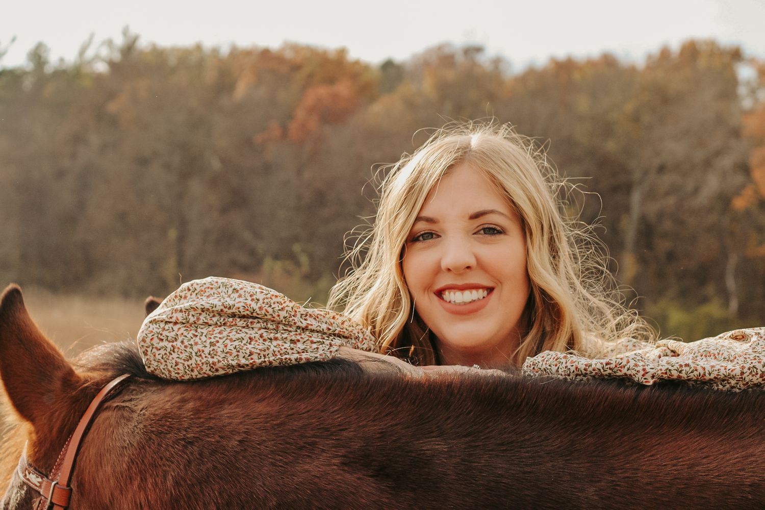 Person resting on chestnut horse's back in autumn field with warm lighting.