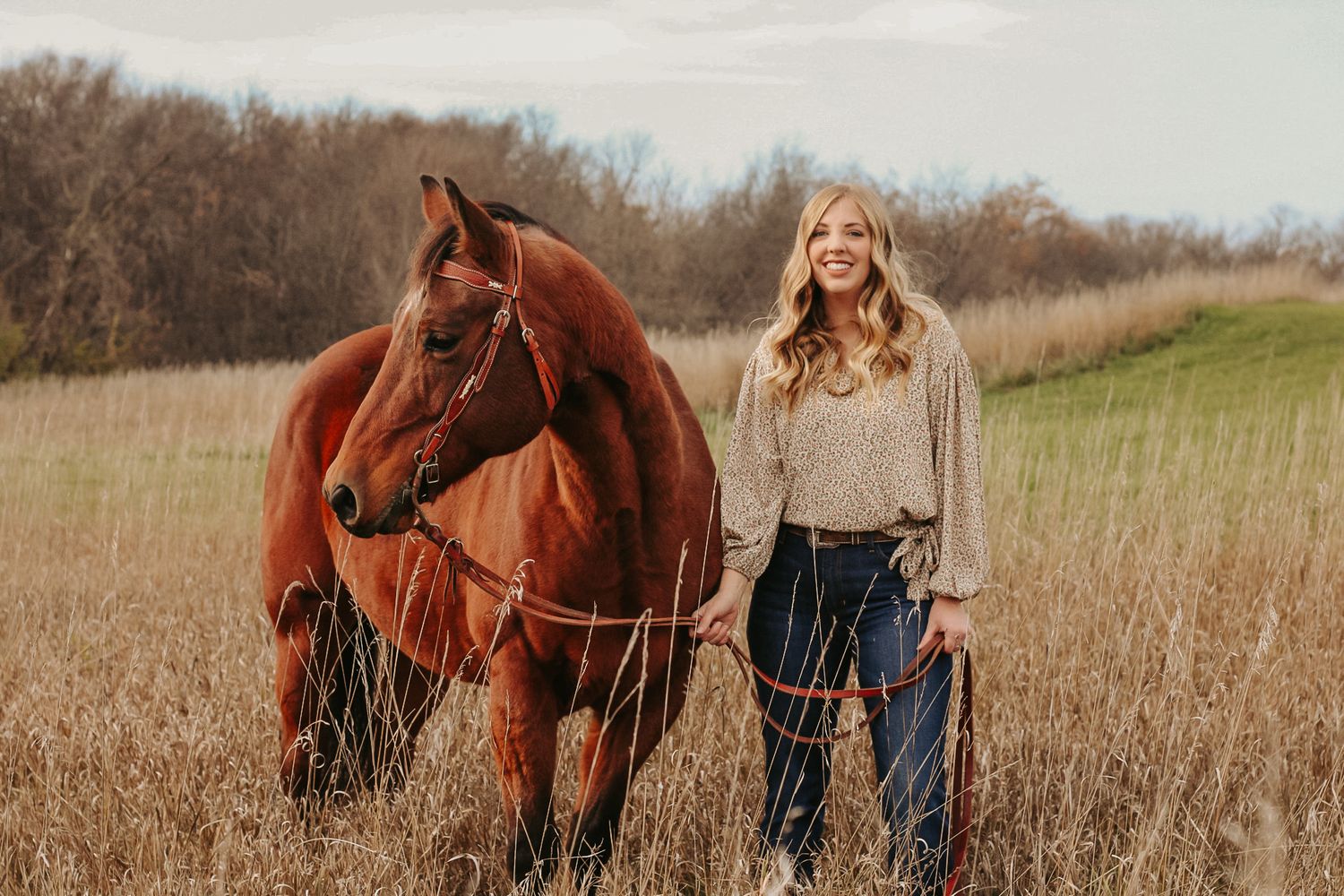 Person in casual attire standing with chestnut horse in tall grass field.