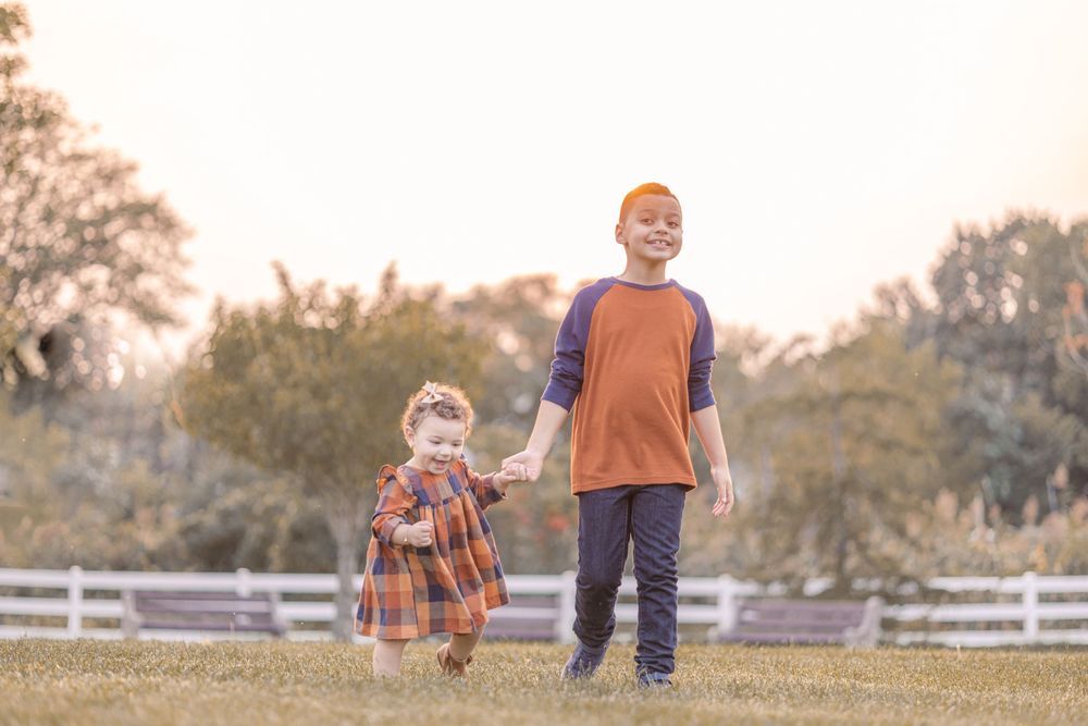 Two children holding hands while walking together in a sunny field with white fence.
